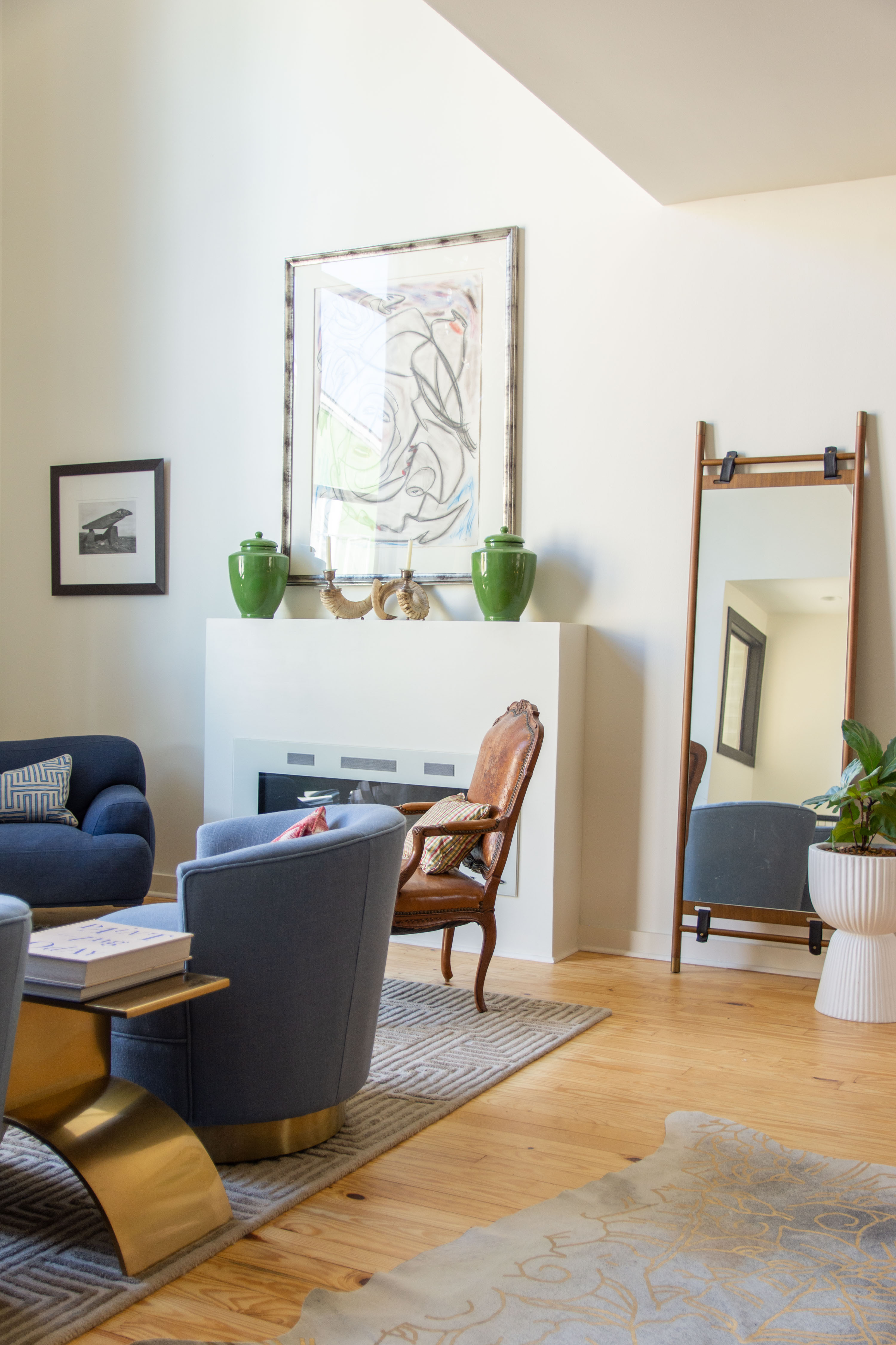 Modern living room design with a cozy yet elevated aesthetic. This space features deep blue accent chairs paired with a gold base coffee table, creating a chic contrast against the neutral backdrop. A statement floor mirror leans against the wall, adding depth and natural light reflection. The fireplace mantle is styled with bold green ceramic vases and sculptural decor, complemented by abstract wall art for a contemporary touch. Warm wood floors and layered area rugs bring in texture, while a mix of vintage and modern seating makes the room inviting. Perfect inspiration for living room styling, modern home decor, and transitional interior design. 

 