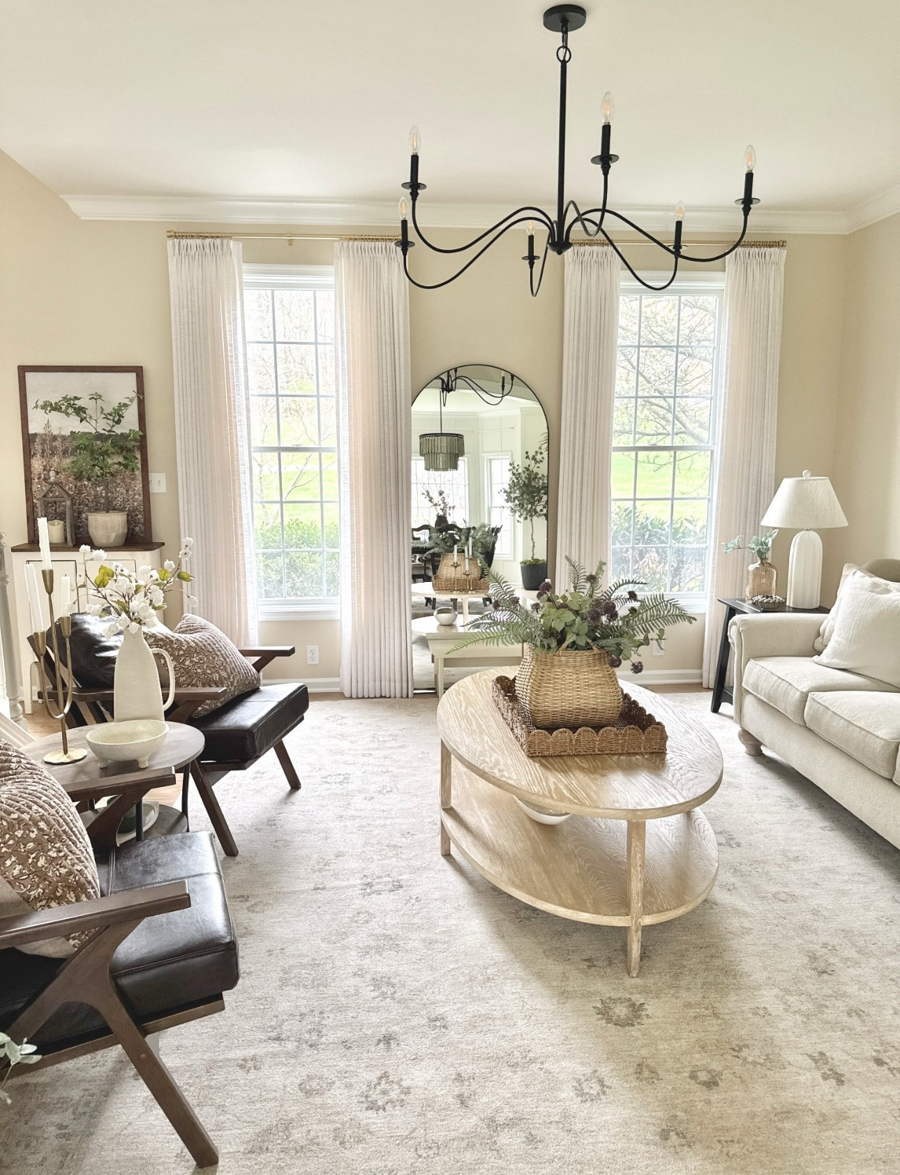 Neutral vintage inspired area rug, curved black metal chandelier, custom curtains and gold hardware, solid wood oval coffee table, faux leather accent chairs, scalloped rectangular tray, woven baskets, faux ferns and stems, large arched floor mirror, metal candelabra, ceramic bowls and multi stacking scalloped bowls  

