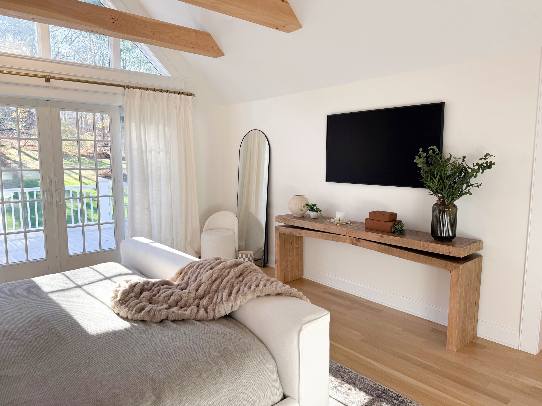 A cozy corner of the bedroom showcases a sleek TV mounted above a stylish console with clean lines and rich wood tones. Beside it, a tall mirror adds depth and reflects natural light, while an accent chair tucked nearby creates the perfect spot for reading or relaxing. The thoughtful layering of textures and neutral palette ties everything together. 
