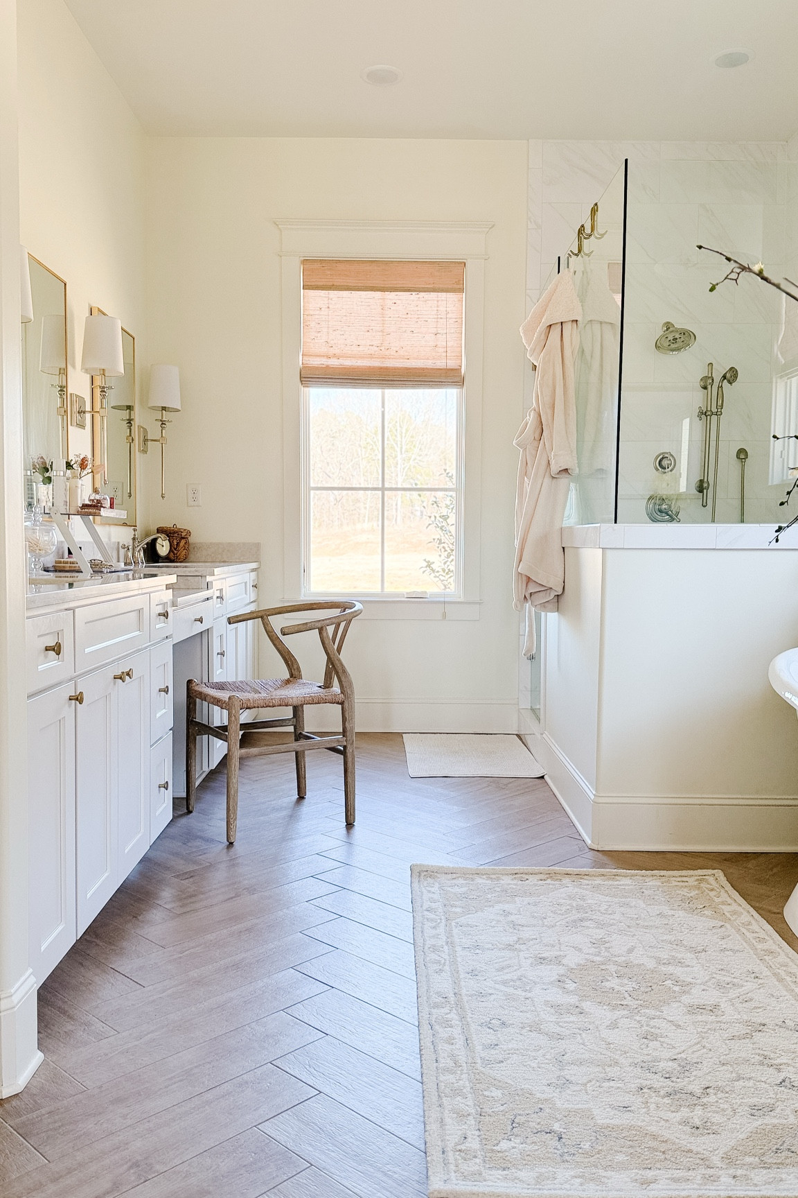 Primary Bathroom Details & What I’d Do Differently 🤍
I realized I’ve never shared many stills of our primary bathroom here, so I thought it was time! Whether you’re planning a remodel, starting a new build, or just love home details, I hope these details (and a few lessons learned) are helpful. I remember how overwhelming it was to source everything when we were building—so hopefully, this saves you a little time!
Flooring:
Wood-look tile floors: Daltile Gainswood in Hickory, grout color: “Latte.” I absolutely love this flooring! With so much white in the space, the warmth of the wood-look tile really helps to ground the room.
Vanity & Paint:
Bathroom Vanity: This was custom-made by a local cabinet company and painted in their version of white, which closely matches Benjamin Moore White Dove.
Wall Color: Benjamin Moore White Dove this is one thing I’d change. Without my paint swatches handy, I picked shower tiles that are a crisp white—and they make the walls look off-white in comparison. Lesson learned: Always take your paint samples with you when selecting tiles! 
Shower & Countertops:
Shower Wall Tile: Polished ceramic, 12x24” (brand unknown).
Countertop: LX Houses Soprano Quartz remnant.
Bathtub & Faucet:
Freestanding Tub: Ours is a double slipper bathtub from Signature Hardware, but it’s been discontinued. A similar option is the Rosalind Soaking Tub. Ours is 72”, and honestly, it’s a bit too long for me (I’m 5’5” and tend to slide down in it). If I could do it over, I’d choose a smaller size.
Tub Faucet: Signature Hardware Deck Mounted Tub Filler Faucet with Cross Handles and Lever Diverter (Includes Hand Shower). The hand shower is the best—it’s perfect for washing my daughter’s hair and makes cleaning the tub so much easier. I use it all the time!
Décor: Mirrors, rug, and decor pieces are linked below. 

#BathroomInspo #HomeRenovationTips #BathroomRemodel #NeutralHomeDecor #ModernTraditionalStyle

#LTKHome