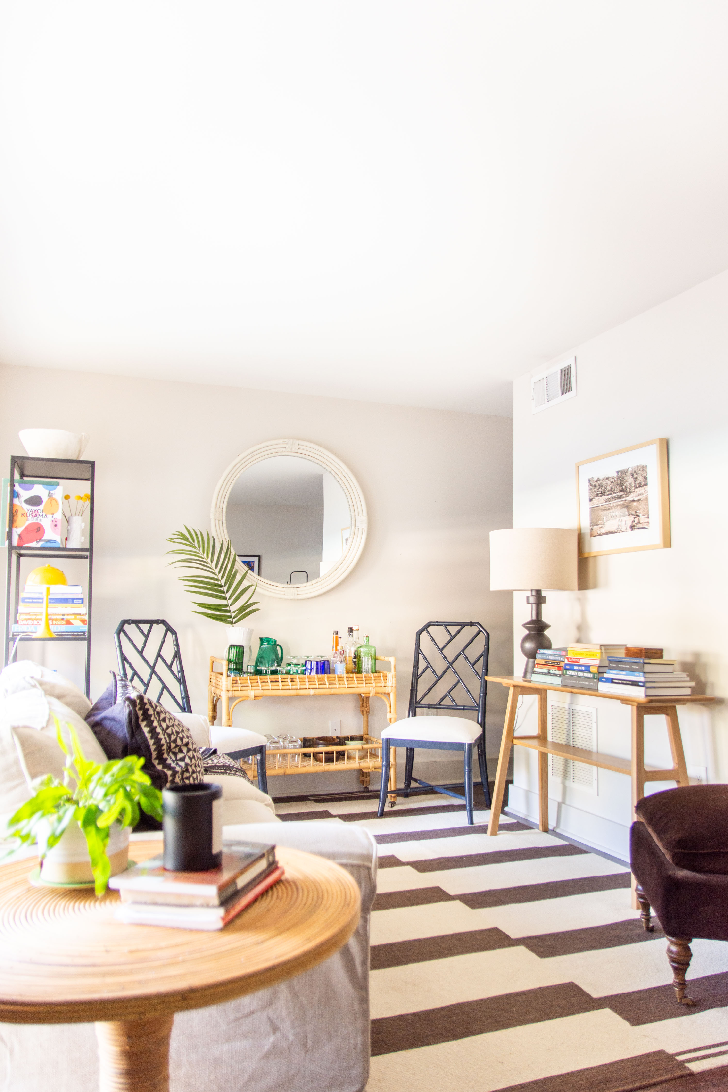Bright and modern living room styling featuring neutral tones with bold accents. A round wood accent table adds warmth, styled with coffee table books and greenery for a touch of freshness. The striped area rug creates visual interest, while the bamboo bar cart with glassware and decor serves as a stylish focal point. Navy dining chairs add contrast against the light walls, complemented by a large round mirror that enhances natural light. A mix of modern and coastal-inspired furniture, layered textures, and curated accessories make this space both inviting and functional. 

 
