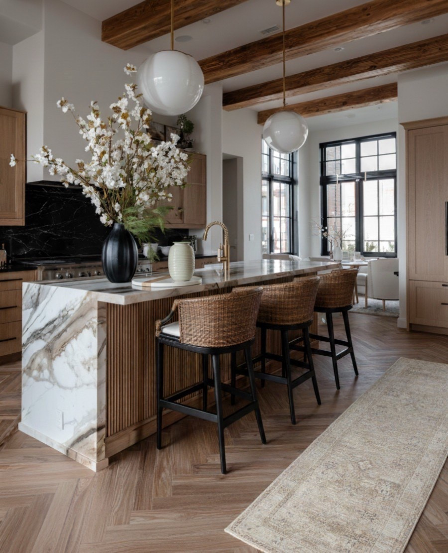 This warm and modern kitchen blends organic textures with timeless design. The waterfall marble island with fluted wood paneling adds natural movement and contrast, complemented by woven rattan counter stools and brushed brass fixtures. Overhead, the globe glass pendant lights bring a soft glow against the exposed wood ceiling beams, tying rustic warmth with elevated modern details. The black marble backsplash, light oak cabinetry, and herringbone hardwood floors complete the layered neutral palette.

 Shop the look:
Waterfall marble island | Fluted wood island paneling | Rattan counter stools | Globe glass pendant lights | Brushed brass kitchen faucet | Black marble backsplash | Light oak cabinetry | Herringbone wood flooring | Neutral vintage runner | Ceramic vases | Faux cherry blossom stems 


#LTKFindsUnder100 #LTKSaleAlert #LTKHome