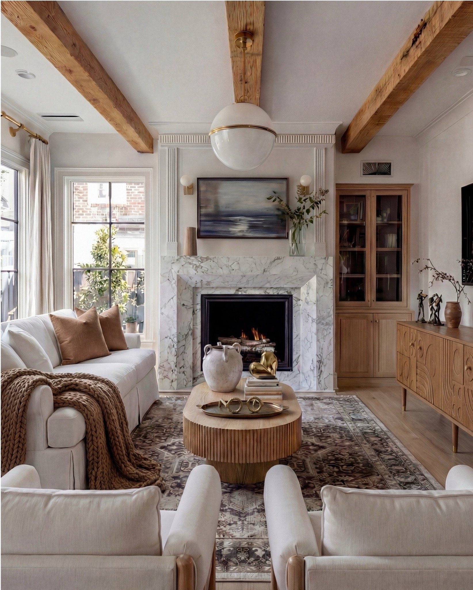 Shop the Look:
• White upholstered sofa (clean-lined, low profile)
• White upholstered armchairs (pair)
• Light wood round coffee table (fluted or ribbed base)
• Neutral vintage-style area rug (muted brown and beige tones)
• Marble fireplace surround
• Black fireplace insert
• Large framed landscape wall art (soft blue and neutral tones)
• Brass wall sconces (pair, globe style)
• White globe pendant light with brass detail
• Natural wood ceiling beams
• Light wood flooring

Coffee Table Styling:
• Large white ceramic vase (rustic sculptural style)
• Decorative hardcover books (neutral covers)
• Gold abstract heart sculpture
• Brass oval tray
• Decorative brass object (sculptural accent)

Sofa Styling:
• Neutral throw pillows (warm beige)
• Chunky knit throw blanket (camel or warm brown)

Media Console Area:
• Light wood sideboard / media console (carved front detail)
• Brass cabinet knobs
• Black figurine sculptures (musician figures, set of three)
• Brown ceramic vase
• Dried branches / minimalist floral stems
• Brass decorative tray


#LTKHome #LTKSaleAlert