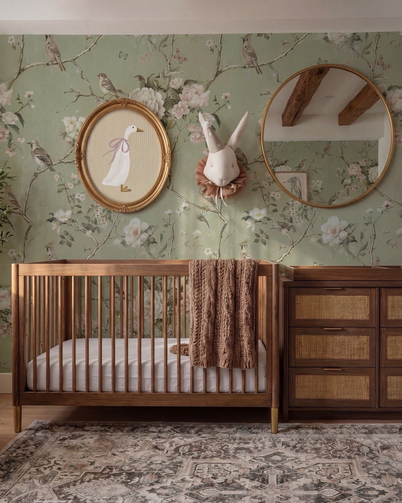This nursery feels storybook-soft and timeless, blending nature-inspired charm with warm, grounding materials. The floral wallpaper sets a whimsical tone, while exposed wood beams and rich wood furniture keep the space feeling elevated and cozy rather than overly sweet.

The balance of classic nursery pieces with refined finishes—like brass accents, woven textures, and sculptural lighting—makes this room feel calm, intentional, and designed to grow with the child.

Shop the Look:
• Floral wallpaper (soft green with botanical print)
• Solid wood crib (warm walnut tone)
• Knit throw blanket (neutral brown)
• Wood dresser with woven drawer fronts
• Changing tray (wood)
• Round brass-framed wall mirror
• Oval framed animal artwork
• Decorative wall animal mount
• Upholstered accent chair (ivory)
• Green accent pillows
• Floor lamp (brass base, white shade)
• Table lamp (ceramic base, white shade)
• Pendant ceiling light (soft white globe)
• Neutral vintage-style area rug
• Light wood flooring
• Sheer curtains with linen drapery panels
#LTKSaleAlert

#LTKsale #LTKhome
