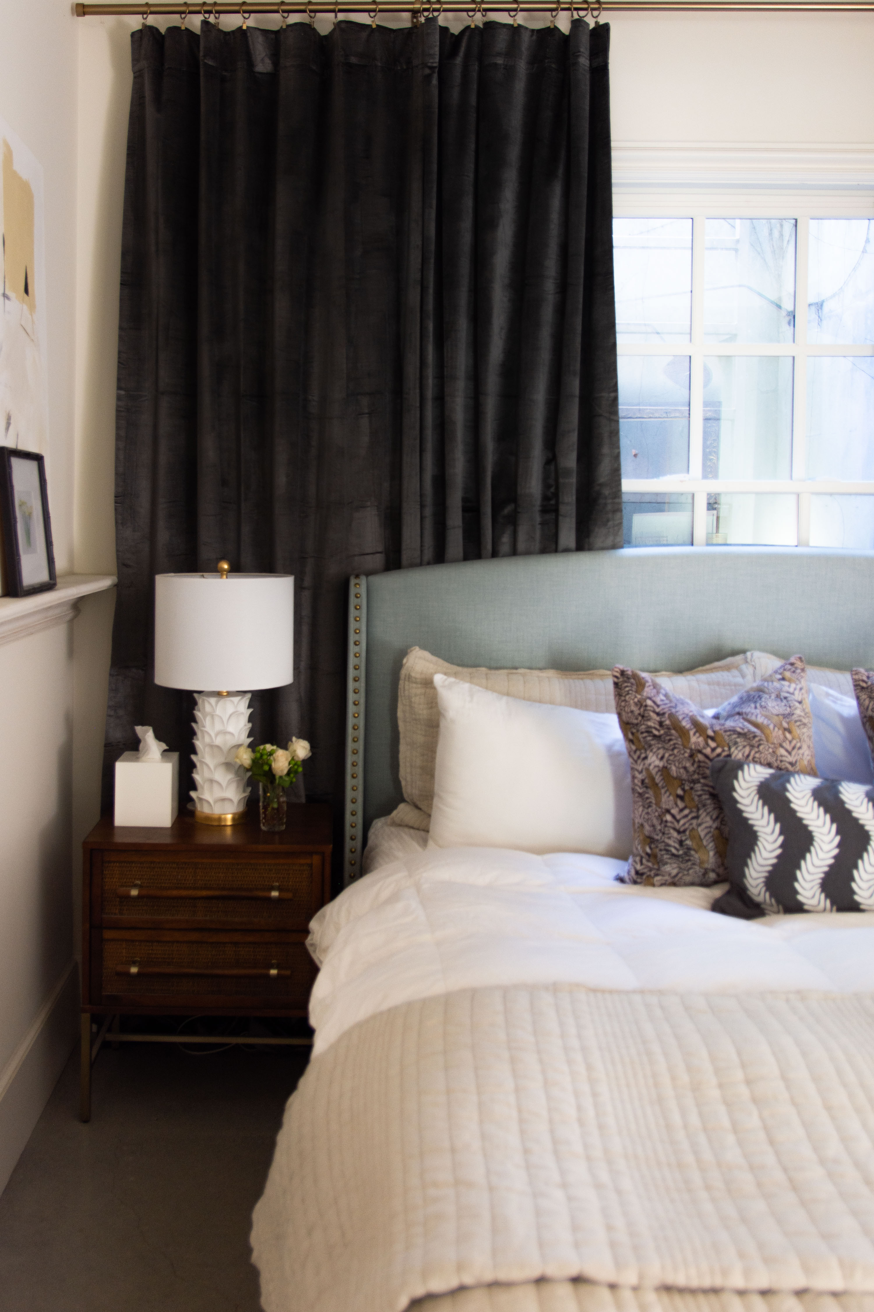 A modern bedroom styled with cozy neutral layers and bold accents. The tufted upholstered headboard in a soft blue-gray anchors the space, paired with crisp white bedding, textured throw pillows, and a quilted beige blanket for warmth. A mid-century nightstand with brass hardware holds a sculptural white lamp, fresh flowers, and simple accessories. Blackout curtains in a rich charcoal tone frame the window, adding contrast and drama. This look blends modern comfort with timeless design, perfect for a chic bedroom makeover. 

 