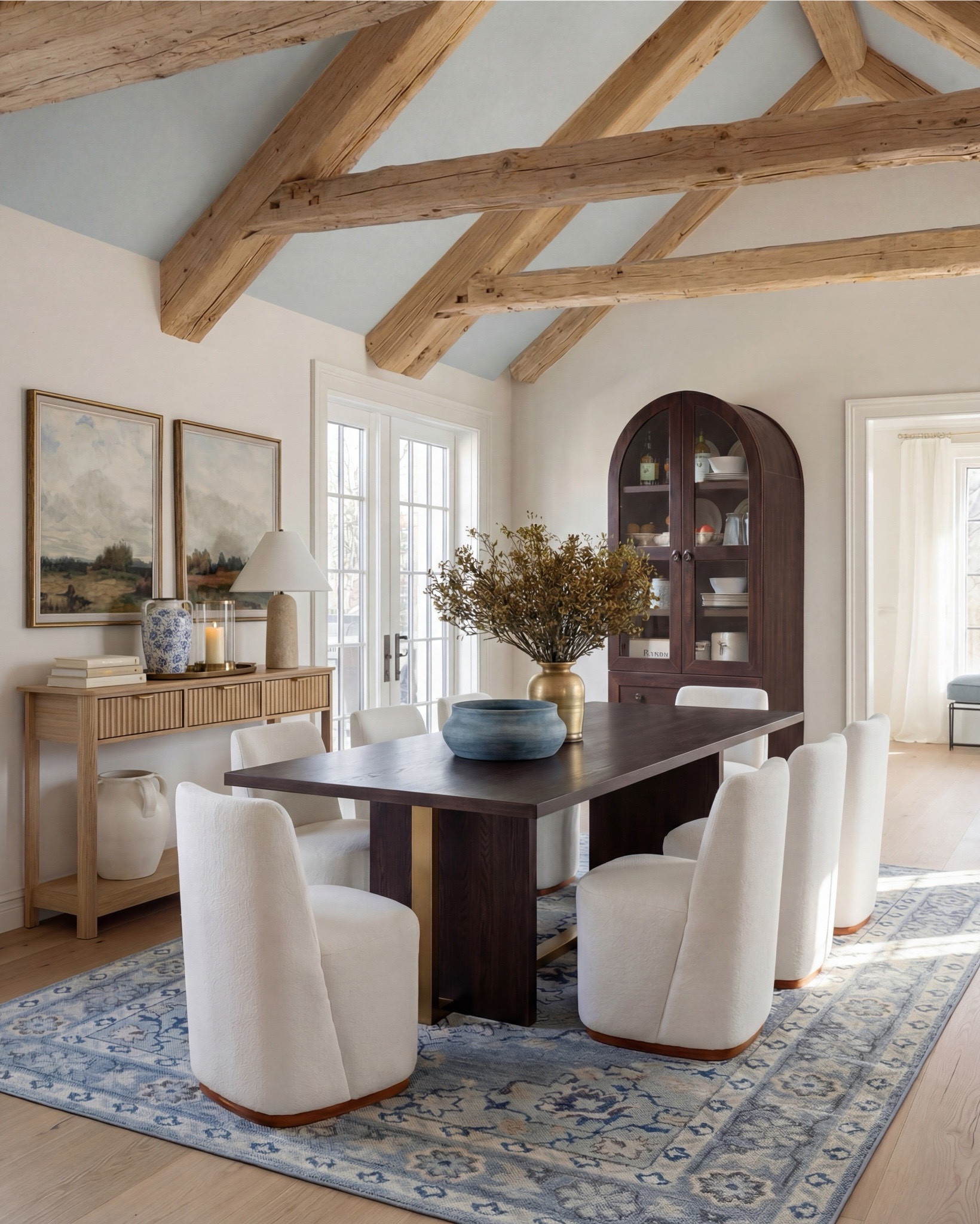 This dining room feels calm, intentional, and quietly elevated. The exposed wood beams warm up the space, while soft blues—on the ceiling and rug—bring in that serene, lived-in charm. The contrast between the dark wood table and the plush ivory chairs makes everything feel grounded yet refined.

The styling really sells it: a blue ceramic bowl centered on the table, a brass vase with organic branches for height, and layered decor along the sideboard that keeps the room from feeling too formal. It’s traditional at its core, but styled in a way that feels fresh and current.

Shop the Look:
• Dark wood rectangular dining table
• Curved ivory upholstered dining chairs (wood base)
• Blue ceramic decorative bowl (table centerpiece)
• Brass vase with dried branches
• Blue patterned area rug
• Light wood sideboard with fluted drawer detail
• Blue-and-white ceramic vase (sideboard)
• Neutral table lamp with white shade
• Stacked books + candle decor
• Framed landscape wall art (set of two)
• Dark wood arched china cabinet with glass doors
• White ceramic floor vase
• Exposed wood ceiling beams
• Soft blue ceiling paint
• Tall floor vase

#LTKsale #LTKhome #LTKstyletip