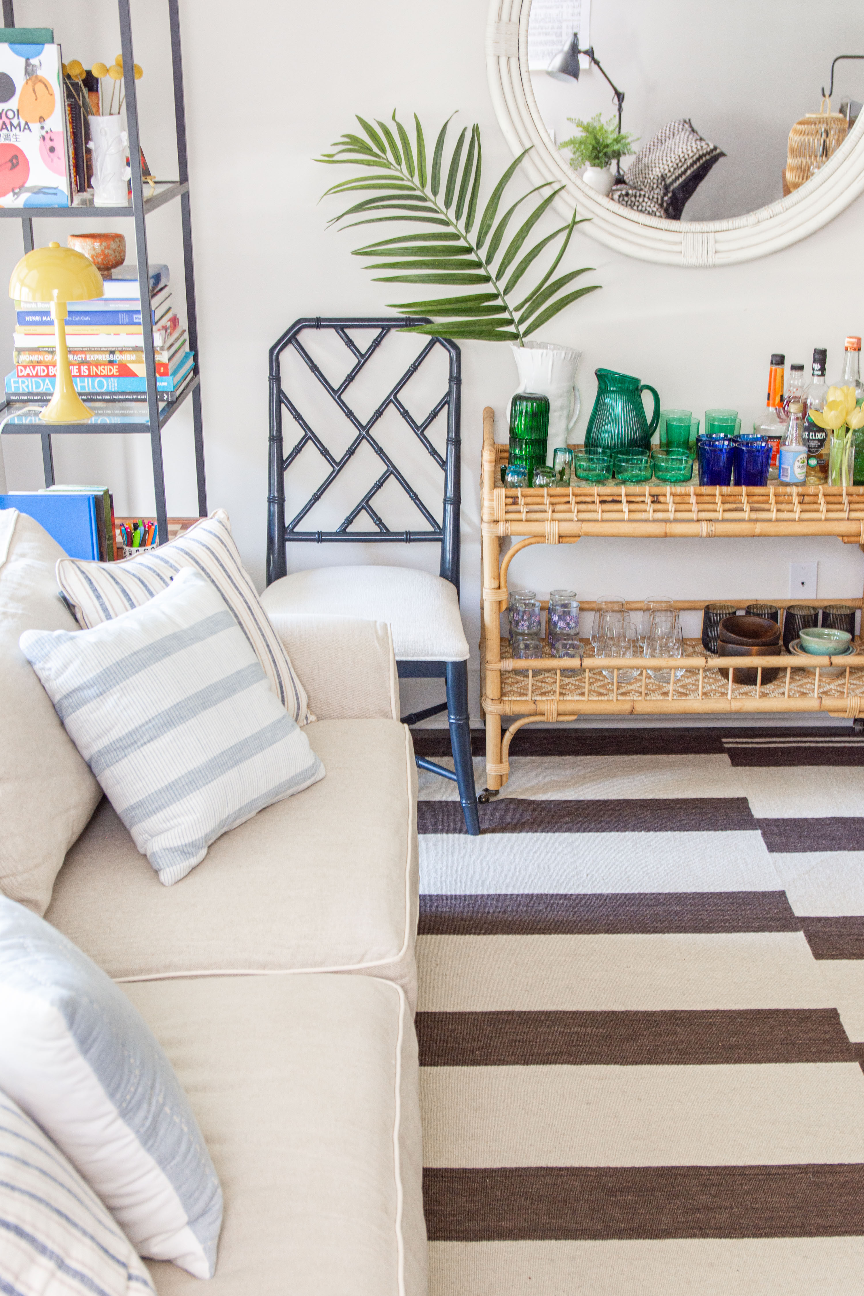 Coastal living meets modern comfort in this bright and inviting space. A neutral linen sofa with striped pillows sets the stage, while a bold black bamboo chair and rattan bar cart add texture and contrast. The striped area rug anchors the room, and layered decor elements—like a round woven mirror, palm leaf arrangement, and colorful glassware—bring personality and style. Perfect for entertaining, the curated mix of coastal accents, natural materials, and pops of color creates a laid-back yet sophisticated vibe. 

 