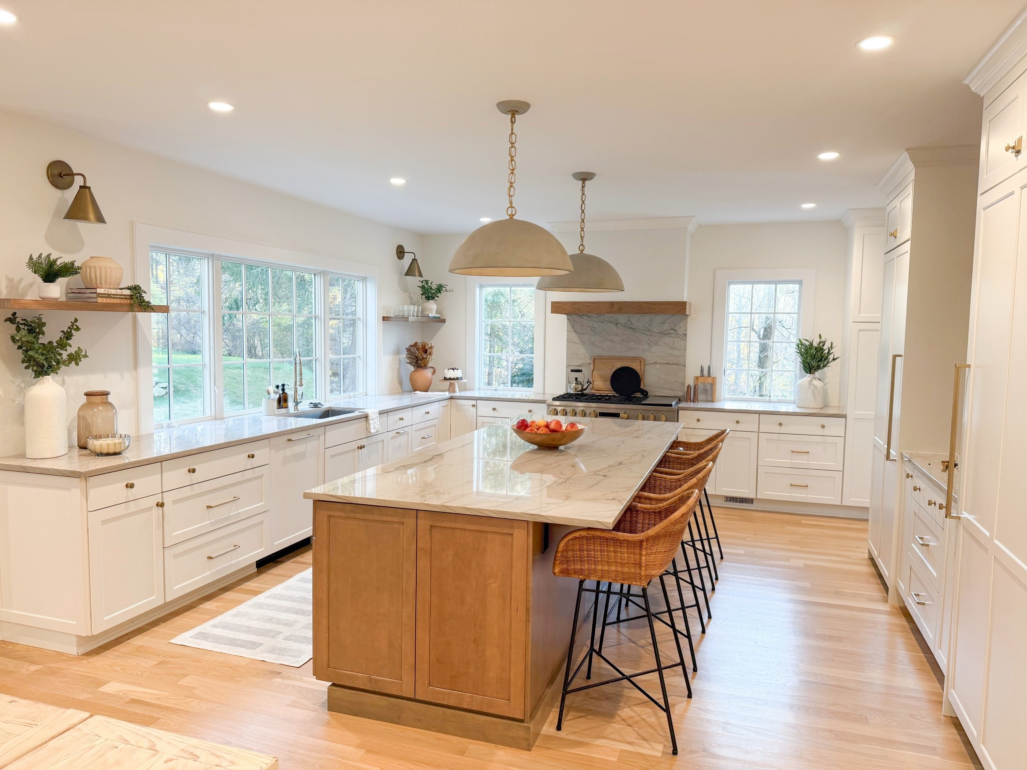 We loved helping our clients with this stunning open-concept kitchen bathed in natural light, showcasing a spacious marble island with a wooden base, rattan barstools, and sleek brass accents. The design blends modern elegance with warmth through white shaker cabinetry, floating wood shelves, and earthy décor elements. The windows bring the outdoors in, making this space both inviting and serene.