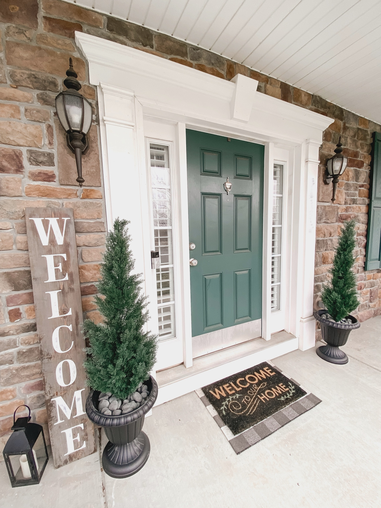 Spring Front Porch Styling
Faux cedar trees in black urn planter with decorative rocks (See below for set up directions!)
Welcome Mat layered with Checkered Rug
Black outdoor lantern
Amazon finds
DIRECTIONS FOR THE PLANTERS:
To make the trees stand securely in the larger planters and also not blow over in the wind:
1. I filled the bottom of the urn planter with the cheap pond pebbles, centered the tree in the middle, and filled in the sides around the tree’s small pot. 
2. Then I added the prettier Mexican Beach pebbles as the top layer.

I used one bag of each type of rock, split between the two planters.

#LTKhome