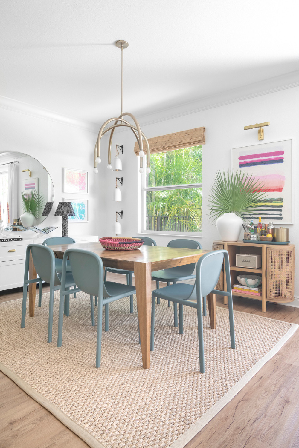 This West Palm Beach Dining room blends coastal and eclectic decor with a natural wood table, muted blue chairs, and a brass-accented chandelier for a warm, inviting look. Colorful Otomi textiles and a woven centerpiece add texture and charm, perfect for family dinners or relaxed gatherings. 

 #LTKHome