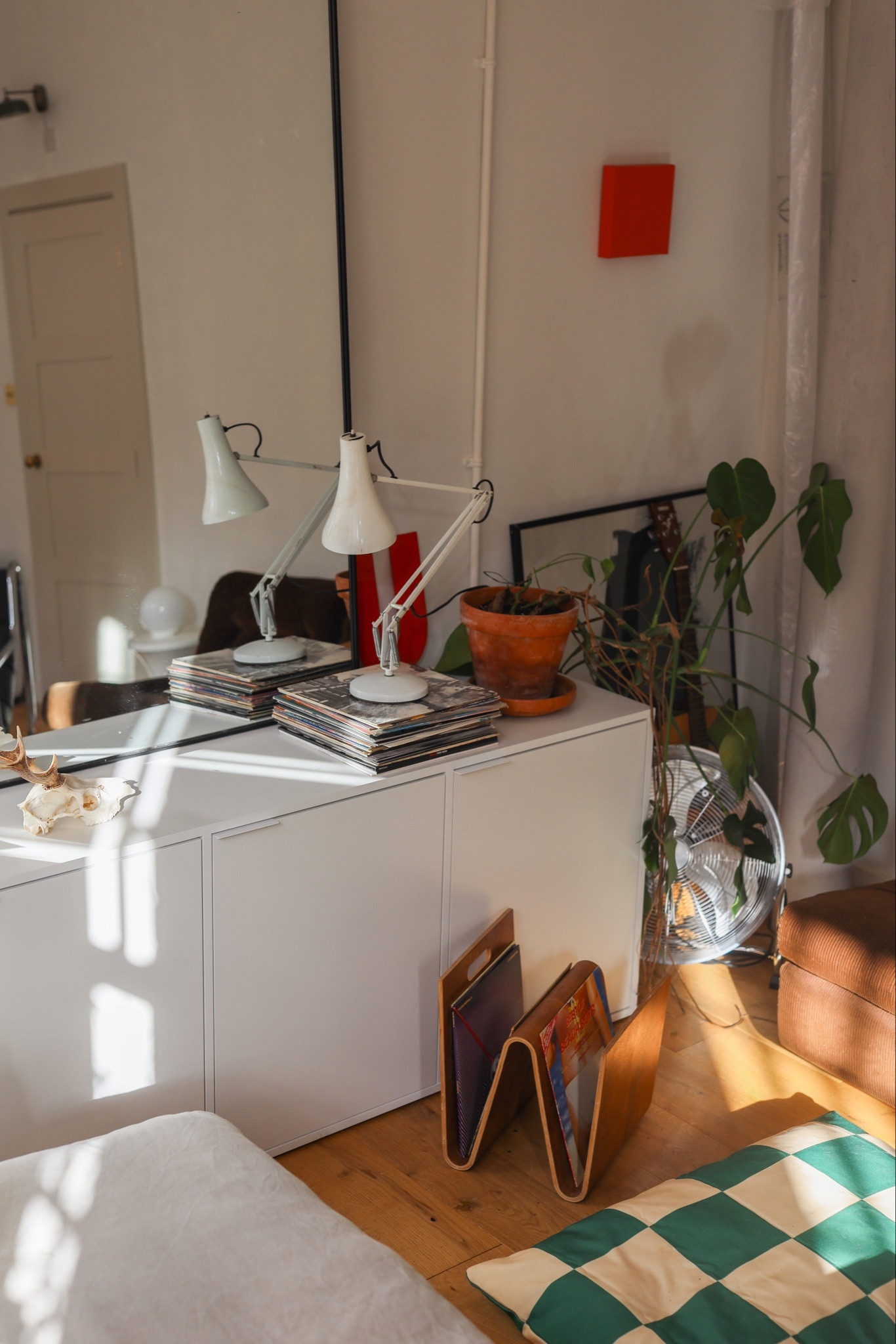 Boho converted factory living room with white minimal Tylko sideboard storage media unit cupboard, monstera houseplant, chrome space age fan, midcentury plywood magazine rack, checkerboard dog bed, vintage anglepoise 90s lamp, stacks of vinyl records and sunlight. Wood floors white walls and high ceilings. Large black mirror. 

#LTKMostLoved #LTKeurope #LTKhome