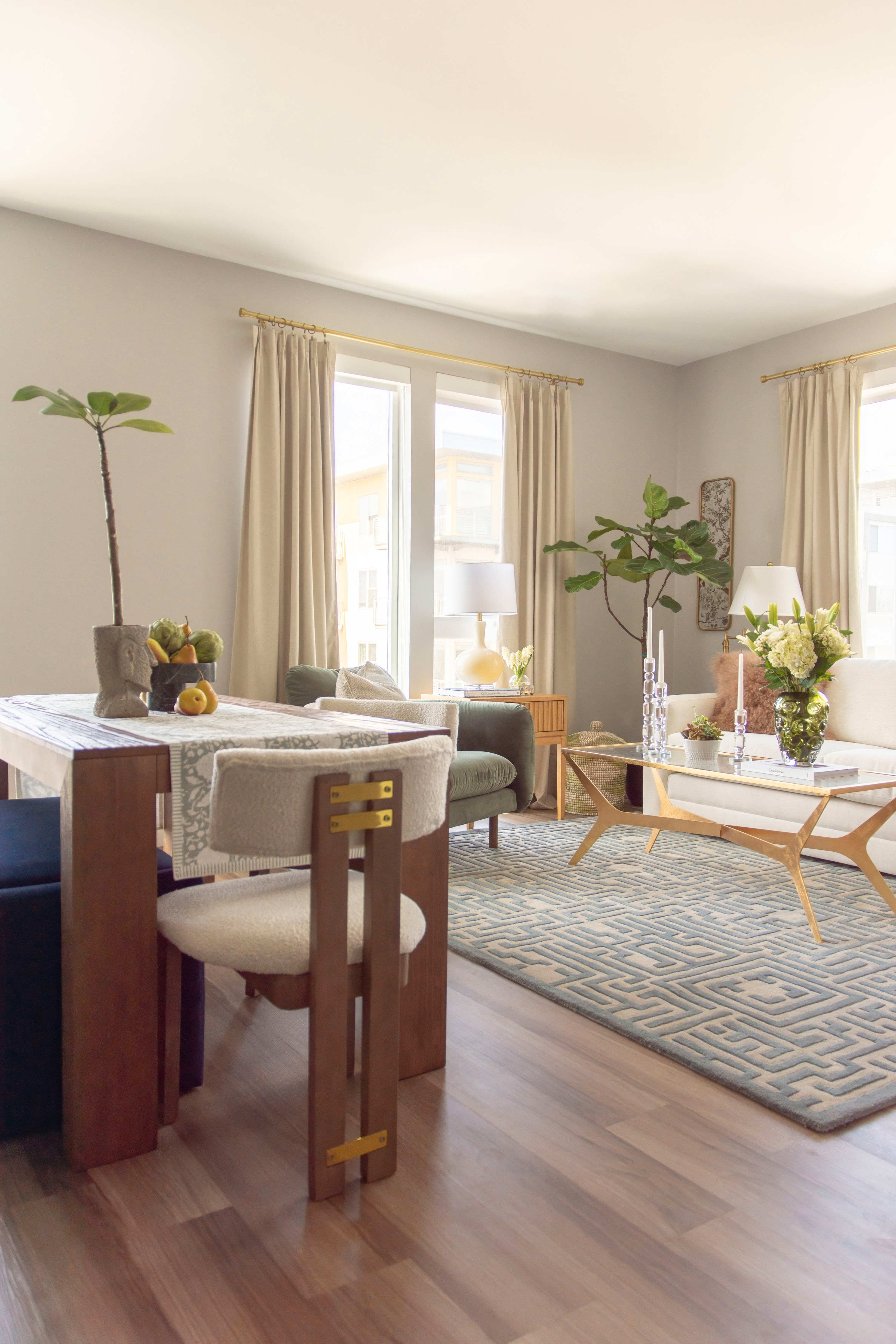 Bright and inviting modern living room design with neutral tones and natural light. This cozy apartment decor features a white sofa, green velvet armchair, mid-century style coffee table with gold accents, and a geometric area rug that anchors the space. Warm wood furniture and boucle dining chairs add texture, while fiddle leaf fig trees and fresh florals bring in organic touches. Perfect inspiration for anyone looking for apartment decorating ideas, neutral home decor, or modern living room styling. 

 