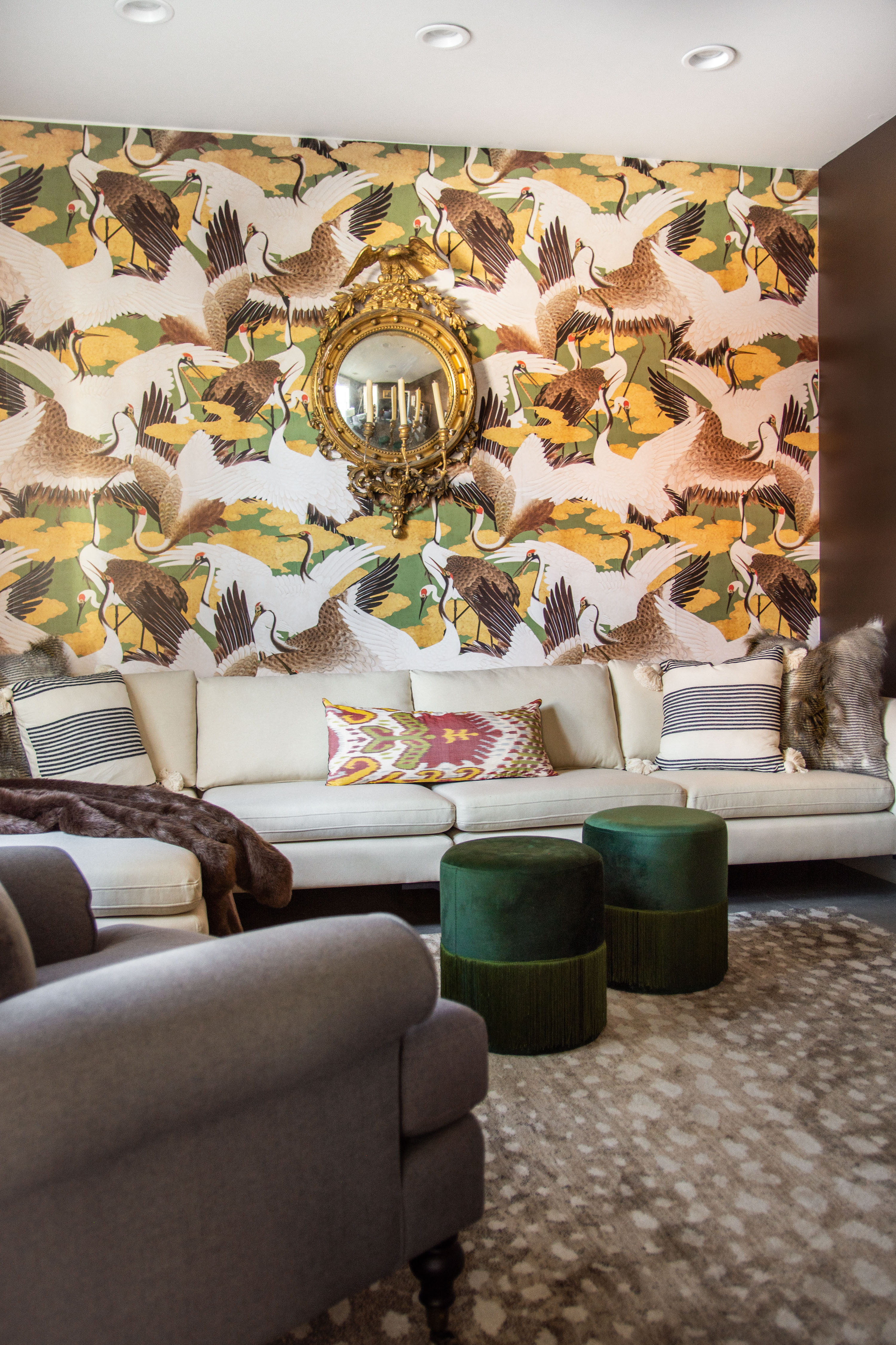 Modern eclectic living room design featuring bold statement wallpaper with cranes, a vintage gold mirror, and a neutral sectional sofa styled with patterned throw pillows. The mix of textures—faux fur throw, velvet green ottomans, and a patterned rug—adds depth and warmth to the space. This cozy yet sophisticated sitting area is perfect for layering neutrals with vibrant accents for a curated home look. Shop statement wallpaper, velvet ottomans, gold mirrors, and neutral sofas to recreate this elevated interior style. 

 