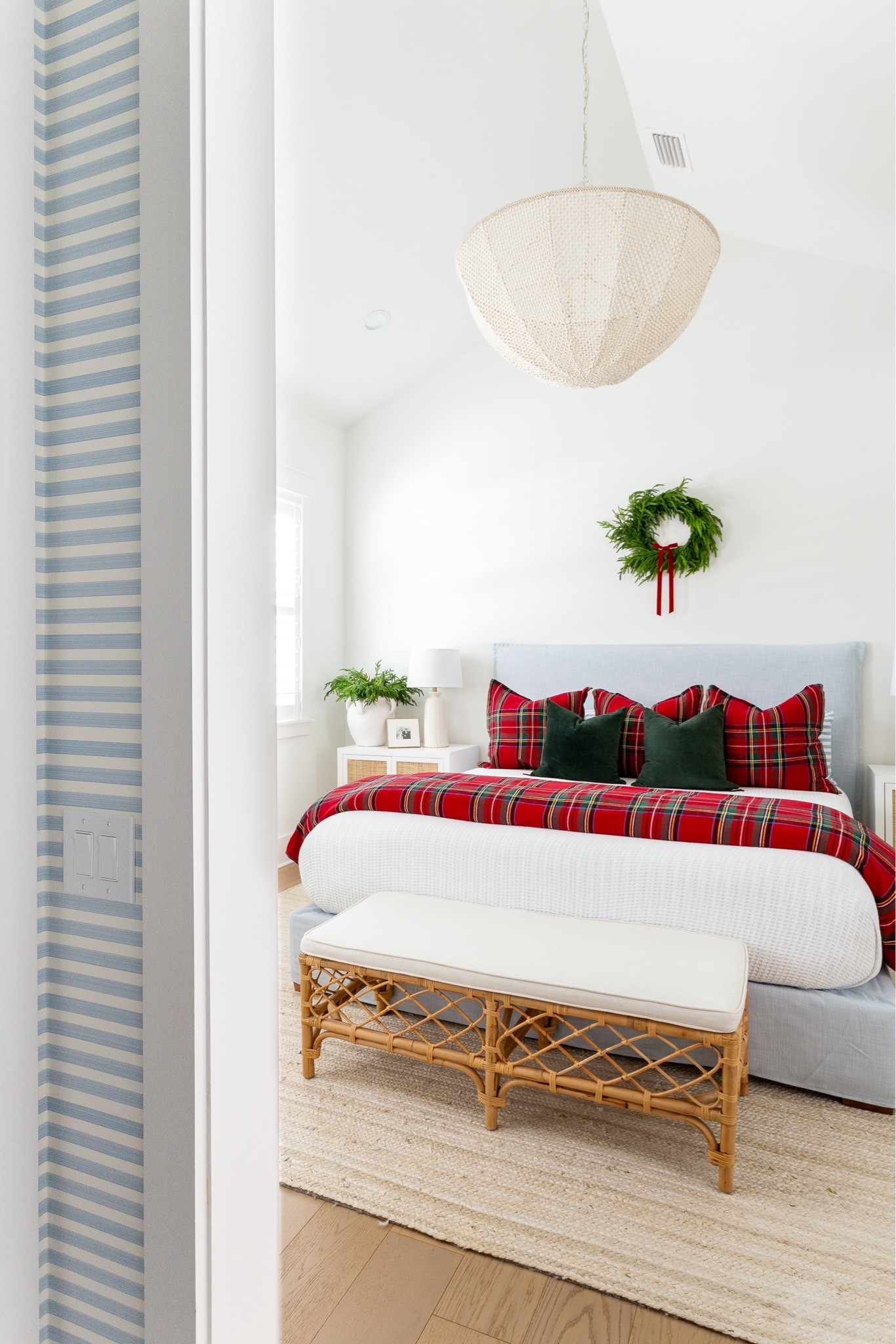A view of our blue striped wallpaper and Christmas bedroom with vaulted ceilings, a light blue linen bed, white and red plaid bedding, dark green velvet pillows, a wreath with red velvet bow above the bed, bead chandelier, rattan nightstands, a rattan bench, and white jute rug. See our full Christmas home tour here: https://lifeonvirginiastreet.com/2024-christmas-home-tour/. #ltkholiday #ltkhome #ltksalealert #ltkfindsunder50 #ltkfindsunder100 #ltkseasonal #ltkstyletip 

#LTKHoliday #LTKHome #LTKSaleAlert