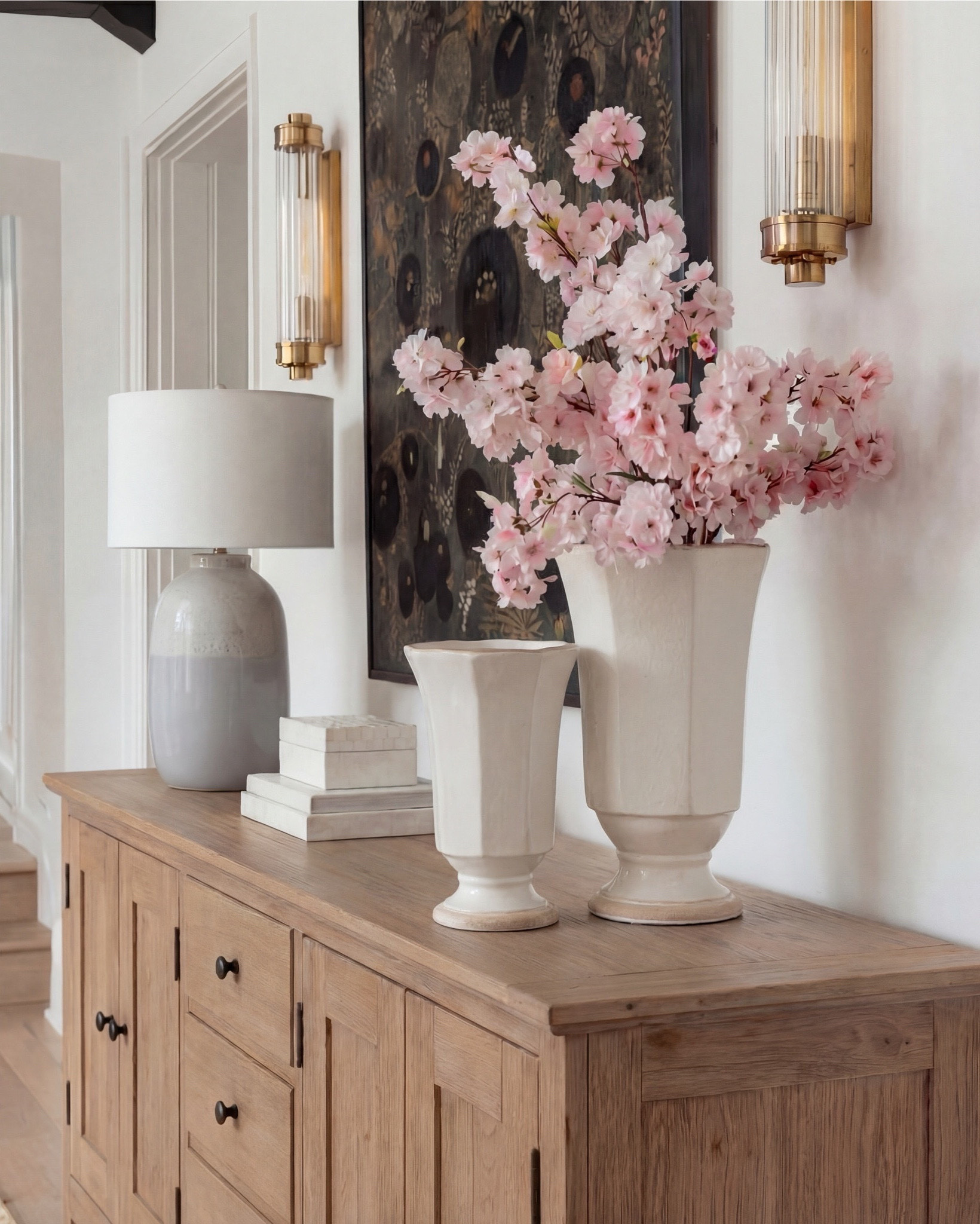 Shop the Look:
• Dark wood pedestal dining table
• Cream upholstered dining chairs with light wood legs
• Large neutral area rug with charcoal medallion pattern
• Black arched display cabinet / hutch
• Light wood sideboard / buffet cabinet
• Large ceramic floor vase (neutral tone)
• White ceramic bowl with white florals (peonies or roses)
• Pink faux cherry blossom stems in white vase
• Neutral ceramic table lamp with white shade
• Tall brass wall sconces with glass detailing
• Framed botanical artwork (set of four)
• Dark abstract wall art (large statement piece)
• White decorative vases (assorted sizes)
• Light linen curtains

#LTKhome #LTKsale #LTKstyletip