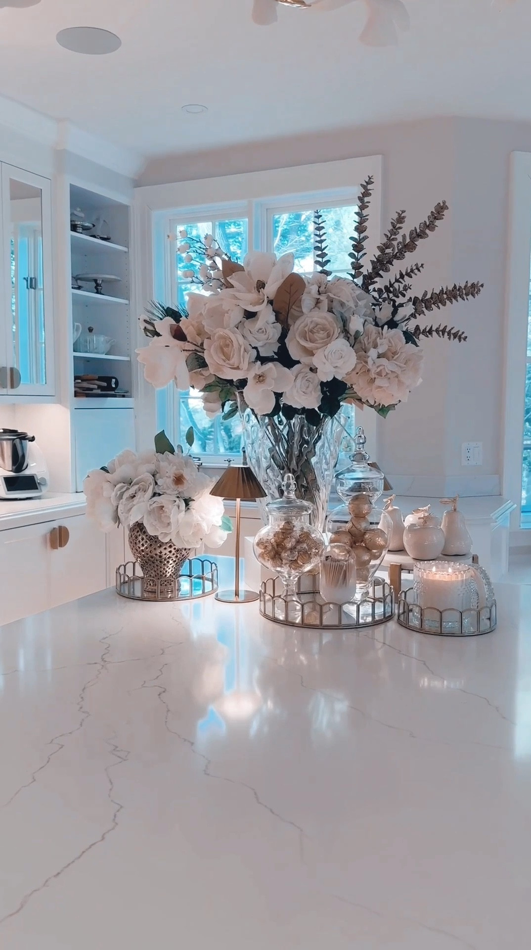 Winter whites, gold accents, and a touch of glamour—this kitchen vignette might just be my favorite yet! ✨ I used mostly sale stems from Christmas (yes, darlings, bargains can be chic!) to create this oversized floral arrangement. With its creamy whites, glittery snowflakes, and sparkling details, it’s the perfect eternal centerpiece. ❄️✨I added scalloped glass trays filled with hot chocolate bombs and gold-wrapped chocolates, complete with apothecary jars for a refined touch. Cream-and-gold apple and pear figurines sit on a marble riser, and a crystal-fresh candle completes the glowing look, especially with the snow outside reflecting light like a scene out of a dream. Stay fabulous and chic! ✨

#LTKFindsUnder50 #LTKHome #LTKSeasonal