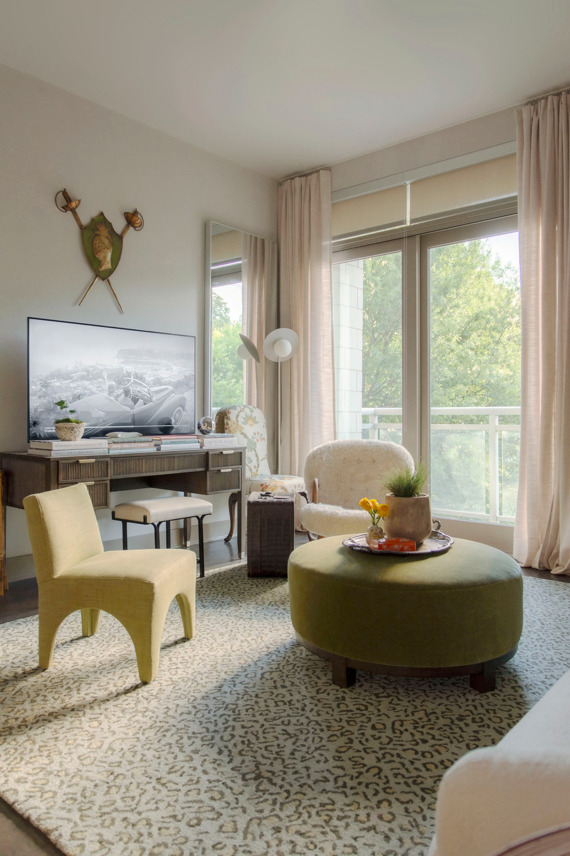 Soft neutrals and layered textures make this cozy living room feel warm and inviting. Sheer blush curtains frame the large windows for an airy look, while a patterned area rug adds subtle dimension. The green velvet ottoman doubles as a coffee table with a chic tray styled with fresh florals and greenery. A boucle accent chair and mid-century inspired side chair bring contrast in shapes and fabrics, while the console table under the TV is styled with stacked books and decorative accents for a polished finish. Perfect inspiration for modern apartment living, small space decorating, and transitional home design. 

 
