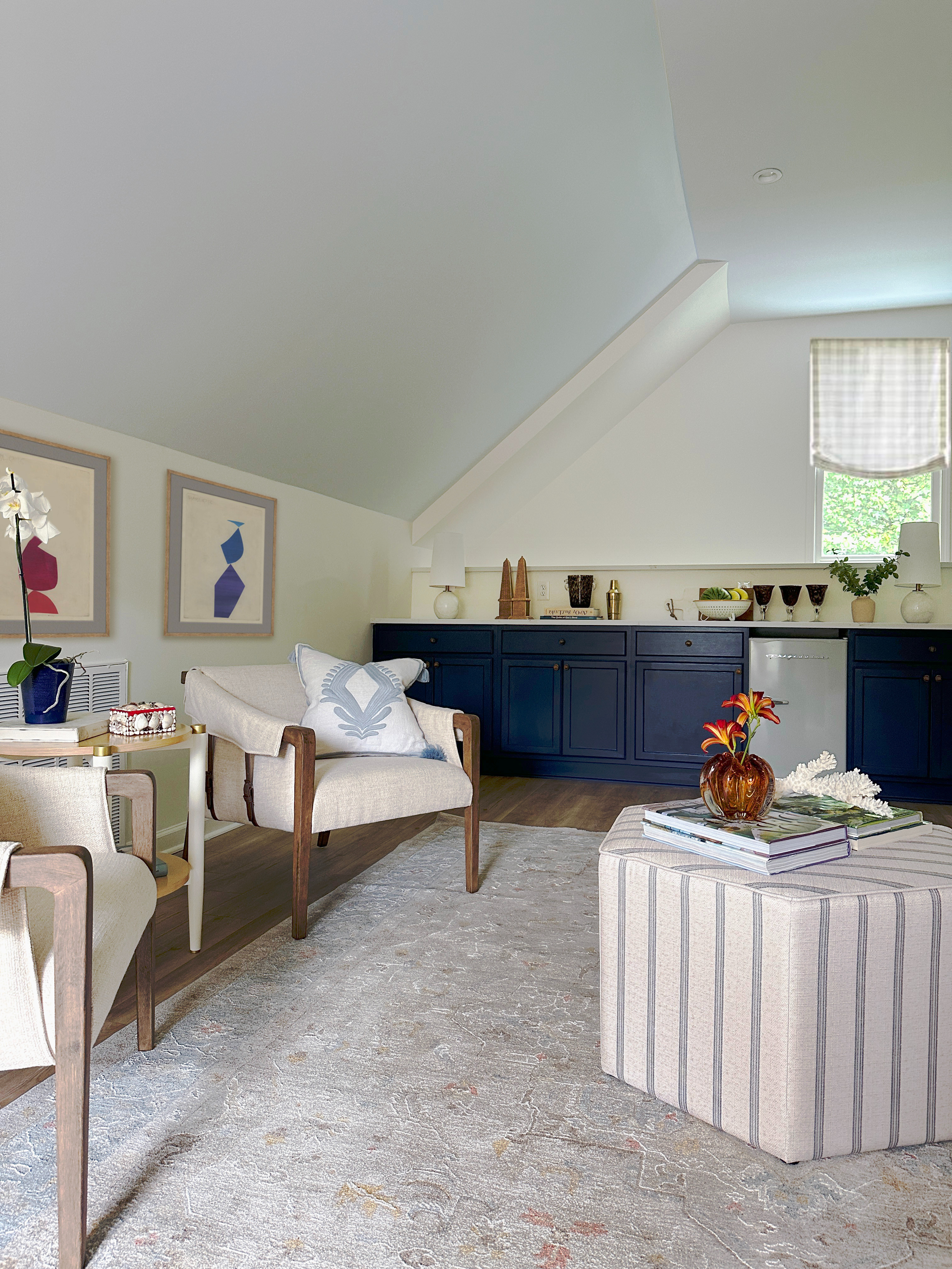 Coastal-inspired guest suite featuring a serene mix of modern and classic design. The space blends neutral tones with pops of deep navy cabinetry, creating a sophisticated yet relaxed atmosphere. A striped ottoman doubles as a coffee table, styled with fresh florals and layered books for an inviting touch. Mid-century modern accent chairs in warm wood and linen bring texture and comfort, while abstract wall art adds a contemporary edge. The soft patterned area rug grounds the room with subtle color and detail, making this the perfect blend of cozy and chic. 

 