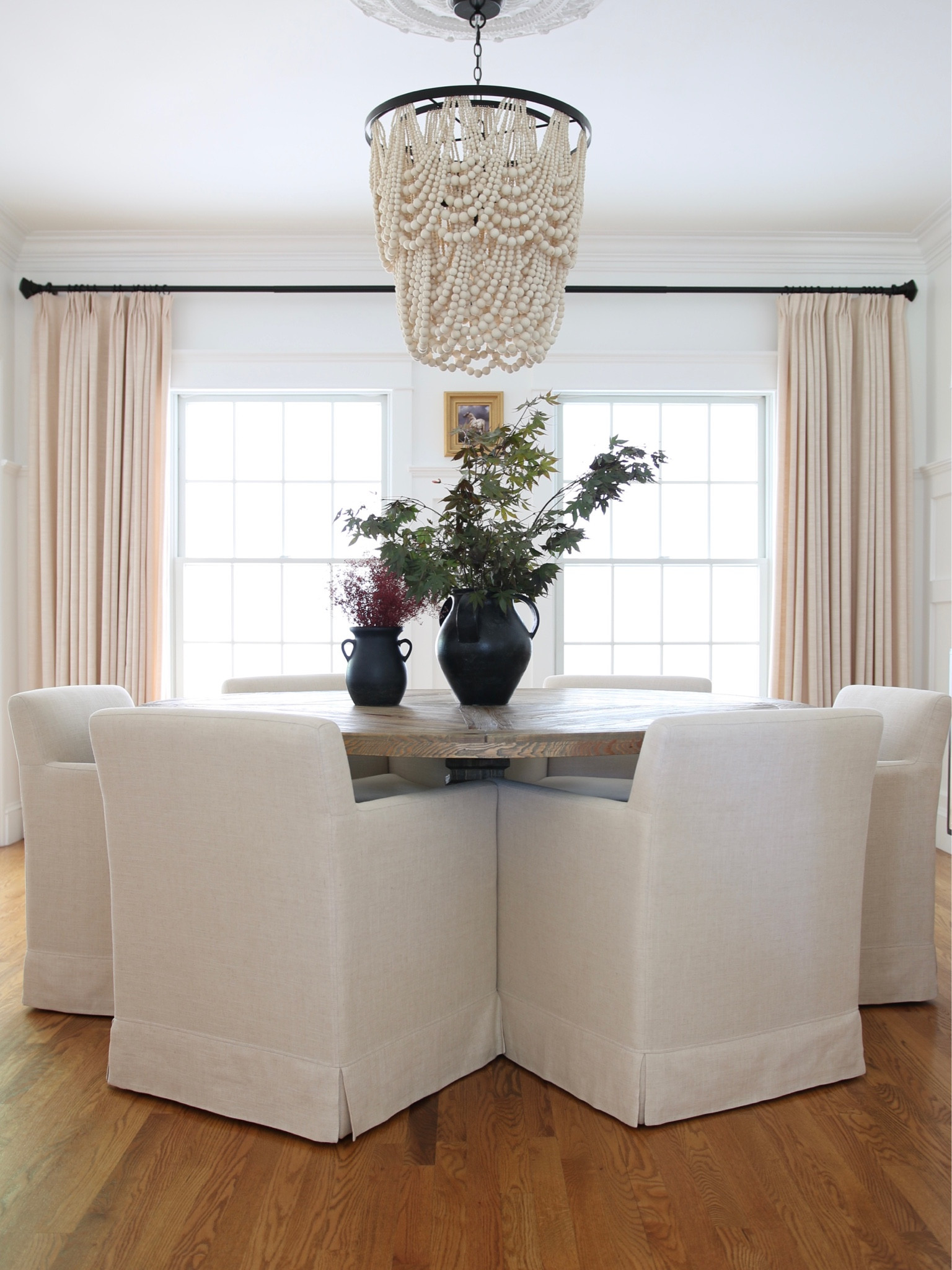 This is my favorite view of my dining room. Originally I had a rectangular table but found that round made more sense. I chose a 71” farmhouse rustic pine pedestal table from Amazon. 
The armchairs are linen upholstered rolling dining chairs from World Market. I have had these for years and they have really held up well. 
I chose Pottery for the ceramic vases, wood bead chandelier and the bronze curtain rings rods and finials. 
Finally, I finished with the famous  Amazon pinch pleated drapes.  

#LTKhome #LTKstyletip #LTKFind