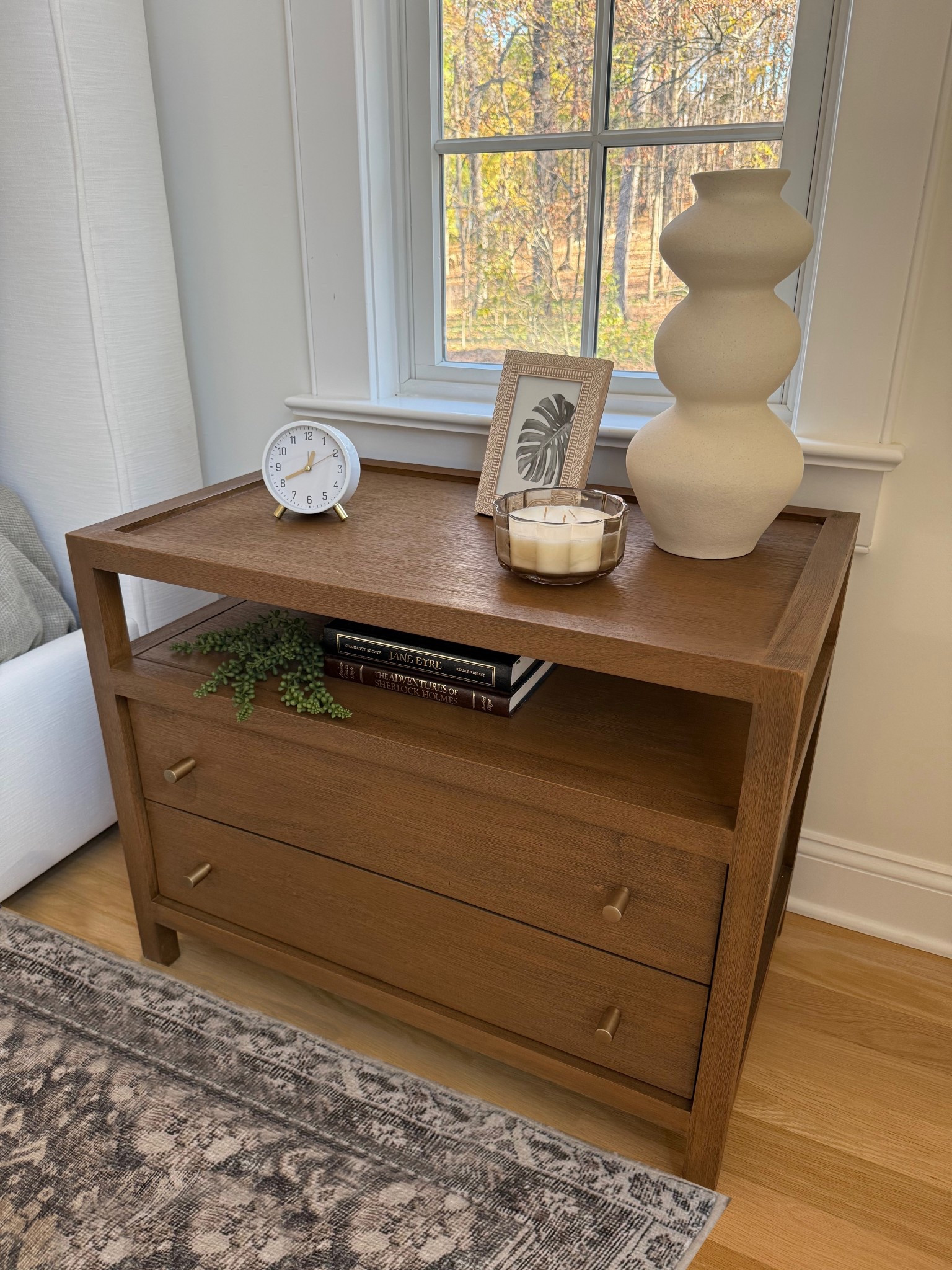 A detailed shot of the beautifully crafted wooden nightstand highlights its rich grain and timeless design. The natural wood finish adds warmth and texture to the space, perfectly complementing the soft tones of the surrounding bedroom decor. The nightstand serves as both a functional piece and a charming accent in this serene primary suite.
