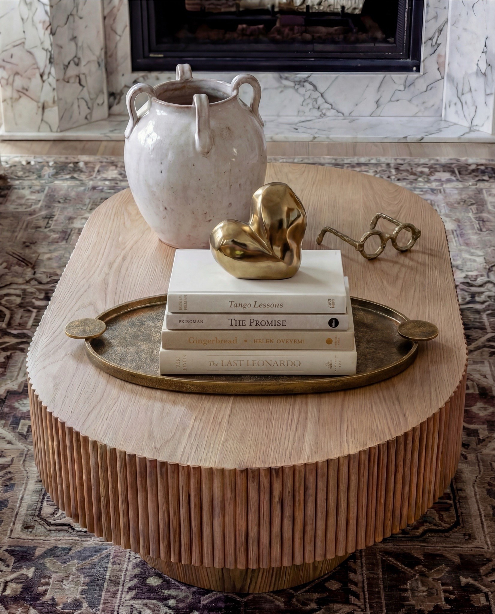 Shop the Look:
• White upholstered sofa (clean-lined, low profile)
• White upholstered armchairs (pair)
• Light wood round coffee table (fluted or ribbed base)
• Neutral vintage-style area rug (muted brown and beige tones)
• Marble fireplace surround
• Black fireplace insert
• Large framed landscape wall art (soft blue and neutral tones)
• Brass wall sconces (pair, globe style)
• White globe pendant light with brass detail
• Natural wood ceiling beams
• Light wood flooring

Coffee Table Styling:
• Large white ceramic vase (rustic sculptural style)
• Decorative hardcover books (neutral covers)
• Gold abstract heart sculpture
• Brass oval tray
• Decorative brass object (sculptural accent)

Sofa Styling:
• Neutral throw pillows (warm beige)
• Chunky knit throw blanket (camel or warm brown)

Media Console Area:
• Light wood sideboard / media console (carved front detail)
• Brass cabinet knobs
• Black figurine sculptures (musician figures, set of three)
• Brown ceramic vase
• Dried branches / minimalist floral stems
• Brass decorative tray

#LTKHome #LTKSaleAlert