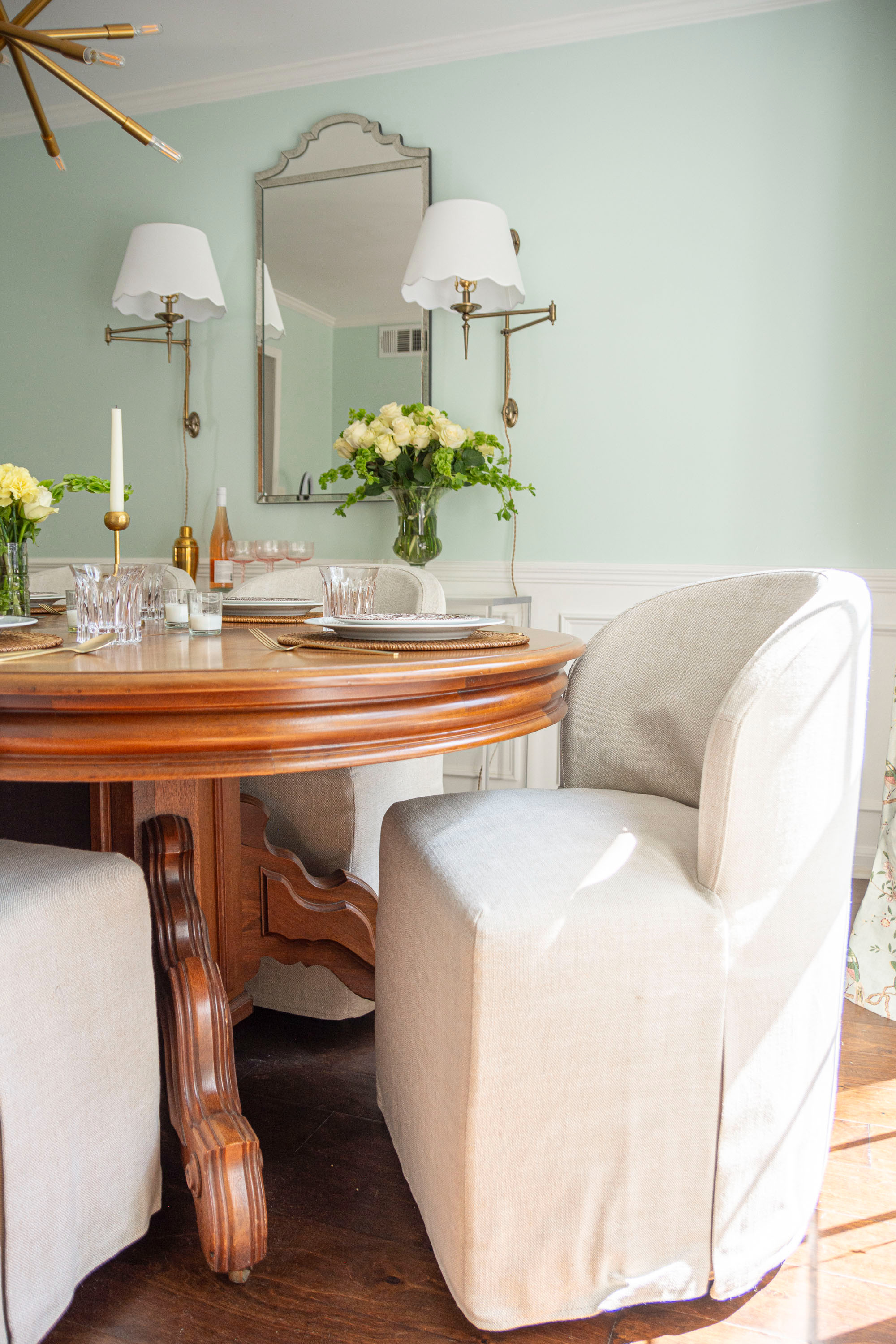 Elegant dining room inspiration featuring a round wooden pedestal dining table paired with slipcovered dining chairs in neutral linen. The space blends traditional and modern style with wall sconces, a statement mirror, and soft mint green walls. Perfect for entertaining, the table is styled with woven placemats, crystal glassware, gold candlesticks, and fresh white roses in a glass vase. This timeless dining room design combines classic furniture, cozy seating, and fresh florals for an inviting yet sophisticated look. 

 