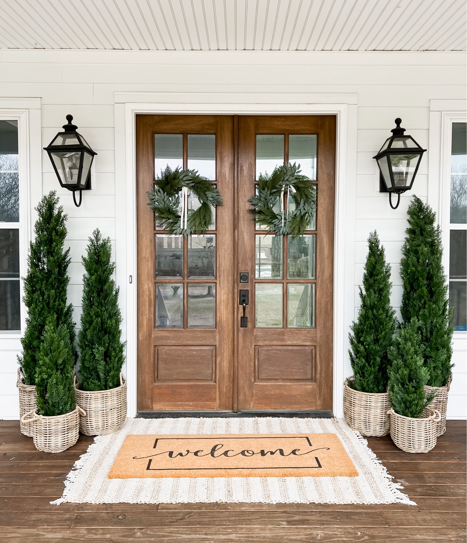 Front porch decor faux artificial cedar trees emerald green arborvitae basket planters double layered doormat and jute, scatter rug, front door and front porch styling wreaths, outdoor wall sconces, lantern lighting modern farmhouse style

#LTKhome #LTKFind #LTKSeasonal