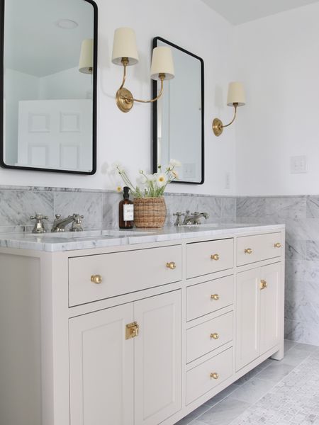 My primary bathroom vanity cabinet came in cashmere grey but I painted it SW accessible grey. I replaced the original hardware with brass ball knobs and latches and added polished nickel faucets. 
Above the vanity cabinet are hand rubbed antique brass single and double face framing scones. Instead of just mirrors I used rounded rectangular recessed medicine cabinets. 
I used Carrara marble for the backsplash, trim and floor  

#LTKFind #LTKstyletip #LTKhome
