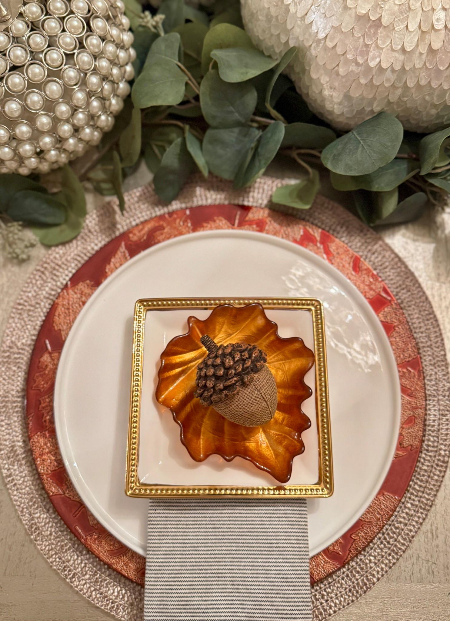 This elegant fall place setting layers warm autumn tones with luxe textures. A woven placemat anchors a red leaf-trimmed charger and crisp white dinner plate, topped with a gilded square appetizer plate. A shimmering amber leaf dish holds a charming burlap-and-pinecone acorn, creating a rustic yet refined seasonal touch. Surrounding pearl and capiz pumpkins with eucalyptus greenery complete the cozy, sophisticated harvest vibe.

#LTKhome #LTKholiday #LTKfall #LTKtabletop #LTKdecor #LTKseasonal #LTKentertaining #LTKinspo #LTKcenterpiece #LTKdining #LTKthanksgiving #LTKstyletip #LTKhomedecor

#LTKSeasonal #LTKStyleTip #LTKHome