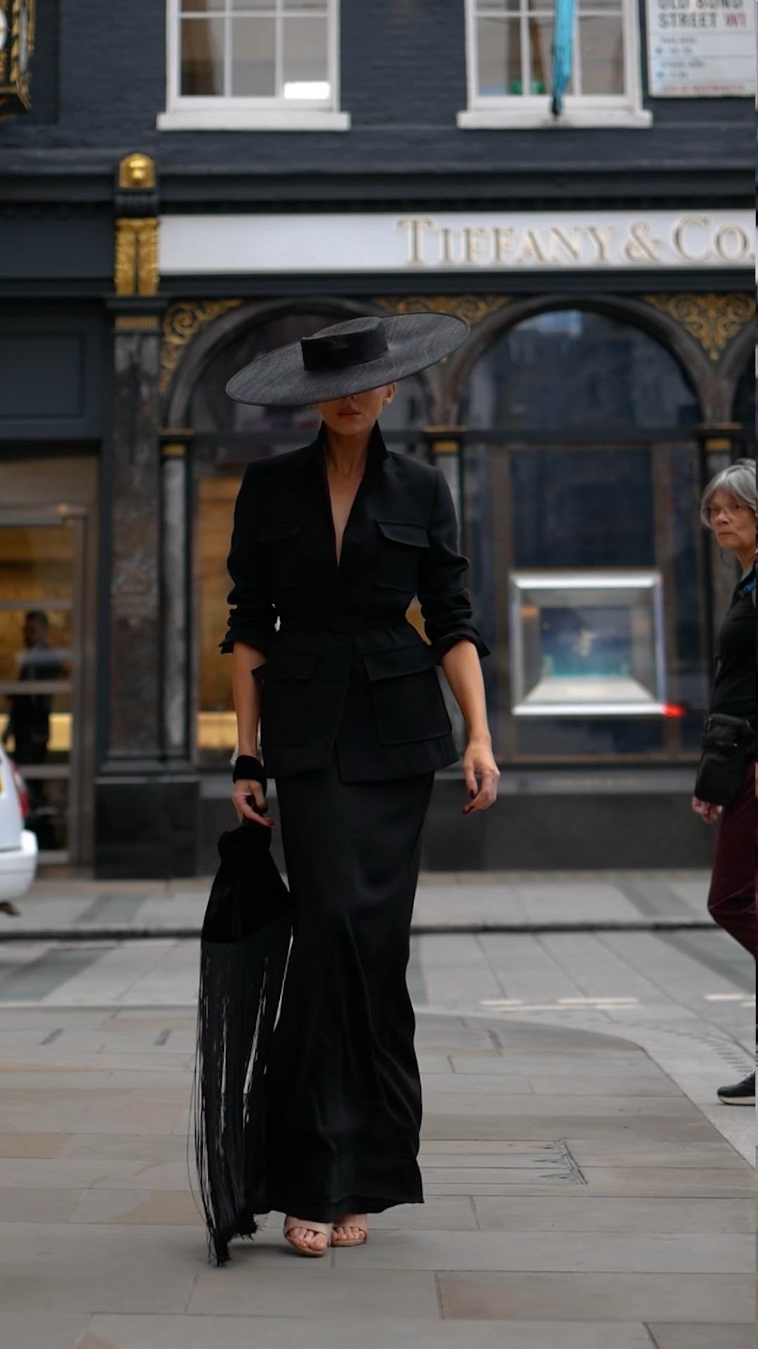 This outfit features a sleek, all-black look with a tailored blazer that cinches at the waist and a long, elegant skirt. The wide-brimmed black hat adds a dramatic touch, while the fringe handbag creates movement and texture. Finishing the look are simple heeled sandals, making for a bold, sophisticated style perfect for formal or fashion-forward occasions.

#LTKluxury #LTKuk #LTKeurope