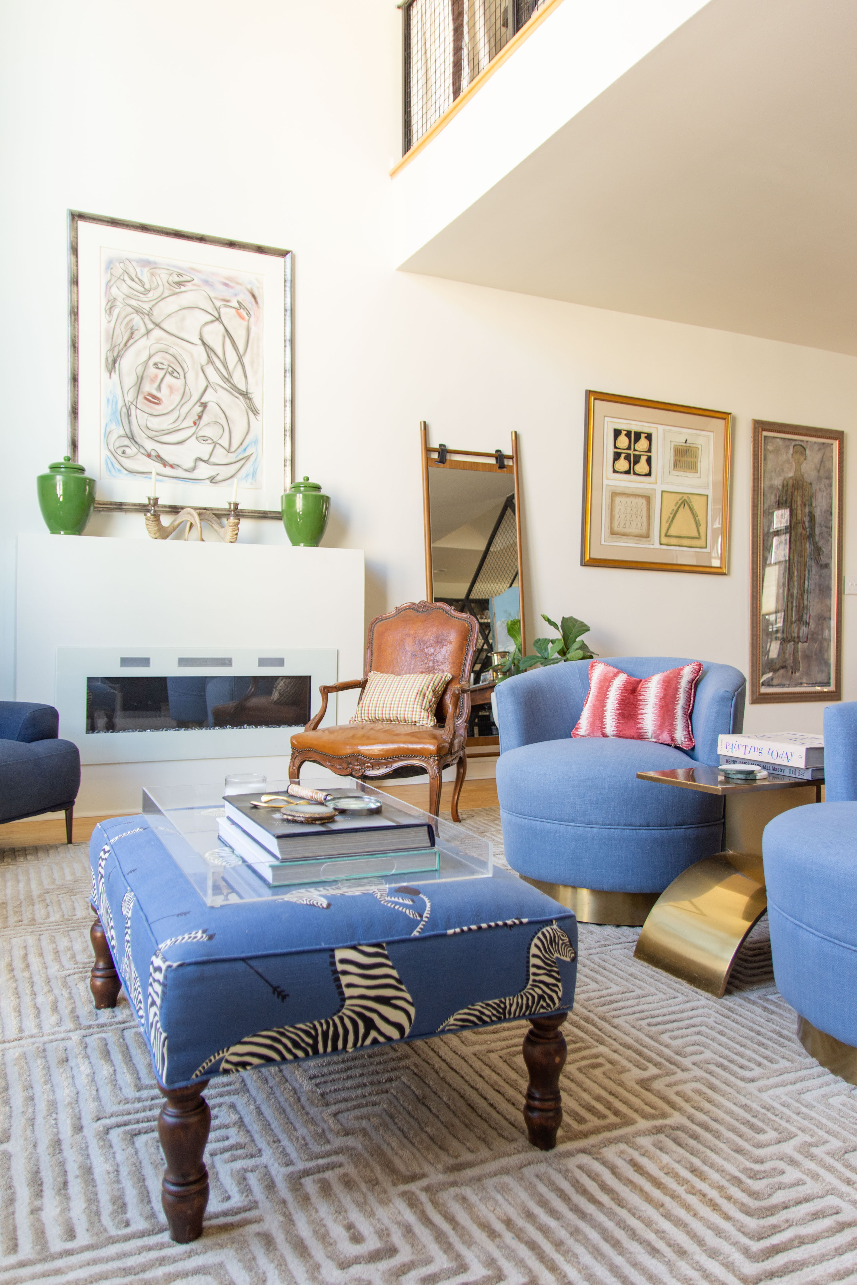 Modern eclectic living room design featuring a mix of classic and contemporary furniture. Bold blue swivel chairs with brass bases pair perfectly with a zebra print upholstered ottoman styled with coffee table books and a clear acrylic tray. A vintage leather accent chair adds warmth and contrast to the neutral geometric rug. The white fireplace surround is topped with green ceramic vases, abstract artwork, and layered wall art for a curated gallery feel. Gold side tables, a leaning mirror, and statement pillows bring extra texture and personality, creating a stylish and cozy space for entertaining or relaxing. 

 
