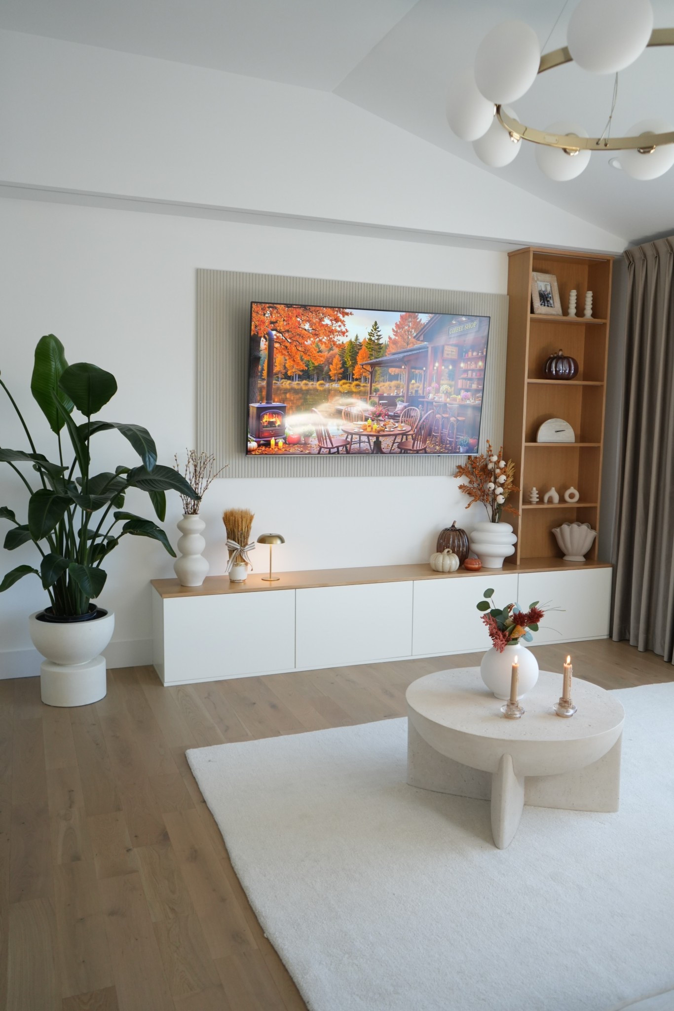 Warm neutrals, cozy textures, and a modern European touch 🤍 This living room setup is all about elevated simplicity — a clean built-in TV wall with fluted detailing, floating media console, and open oak shelving styled with sculptural vases, pumpkins, and fall florals. The round stone coffee table anchors the space perfectly with soft lines and a creamy texture, while the white rug keeps the look bright and airy.

I added two minimalist taper candles and a sculptural vase with fall stems for a cozy seasonal feel

#LTKHome #LTKFindsUnder100 #LTKSaleAlert