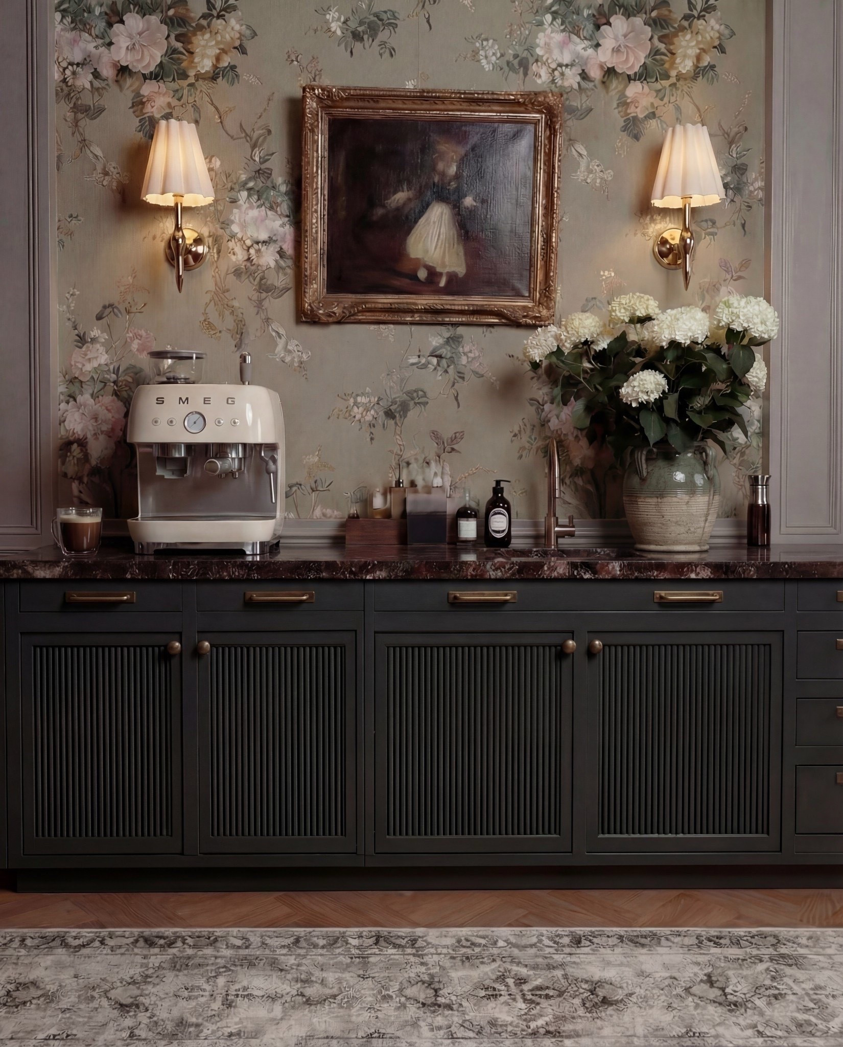 This bar area feels moody, romantic, and old-world refined. Dark cabinetry grounds the space, while floral wallpaper and warm brass accents soften it with a lived-in, European charm. The styling feels curated rather than staged—perfect for slow mornings or evening drinks.

The marble countertop adds quiet luxury, layered with practical bar essentials and sculptural decor. Symmetrical wall sconces frame a vintage-style painting, creating a focal moment that feels intentional and timeless.

Shop the Look:
• Custom bar cabinetry (charcoal or deep green, reeded fronts)
• Brass cabinet pulls and knobs
• Dark marble or stone countertop (brown, burgundy, or black veining)
• Floral wallpaper (muted green, taupe, blush tones)
• Brass wall sconces with pleated fabric shades
• Framed vintage artwork (ornate gold frame)
• Smeg Espresso machine (cream or off-white)
• Ceramic mug or glass cup
• Wooden tray for bar essentials
• Glass bottles and barware
• Stoneware vase (aged or textured finish)
• White hydrangea floral arrangement
• Built-in sink with brass faucet
• Neutral vintage-style runner rug

#LTKOver40 #LTKFestival #LTKHome