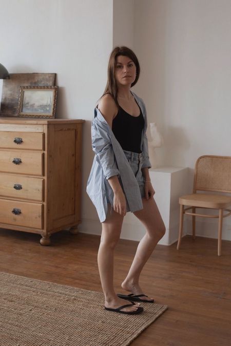 Caroline Joy wears a minimalist summer outfit featuring a black one piece swimsuit with cutoff denim shorts, flip flops, and an oversized button up shirt.
