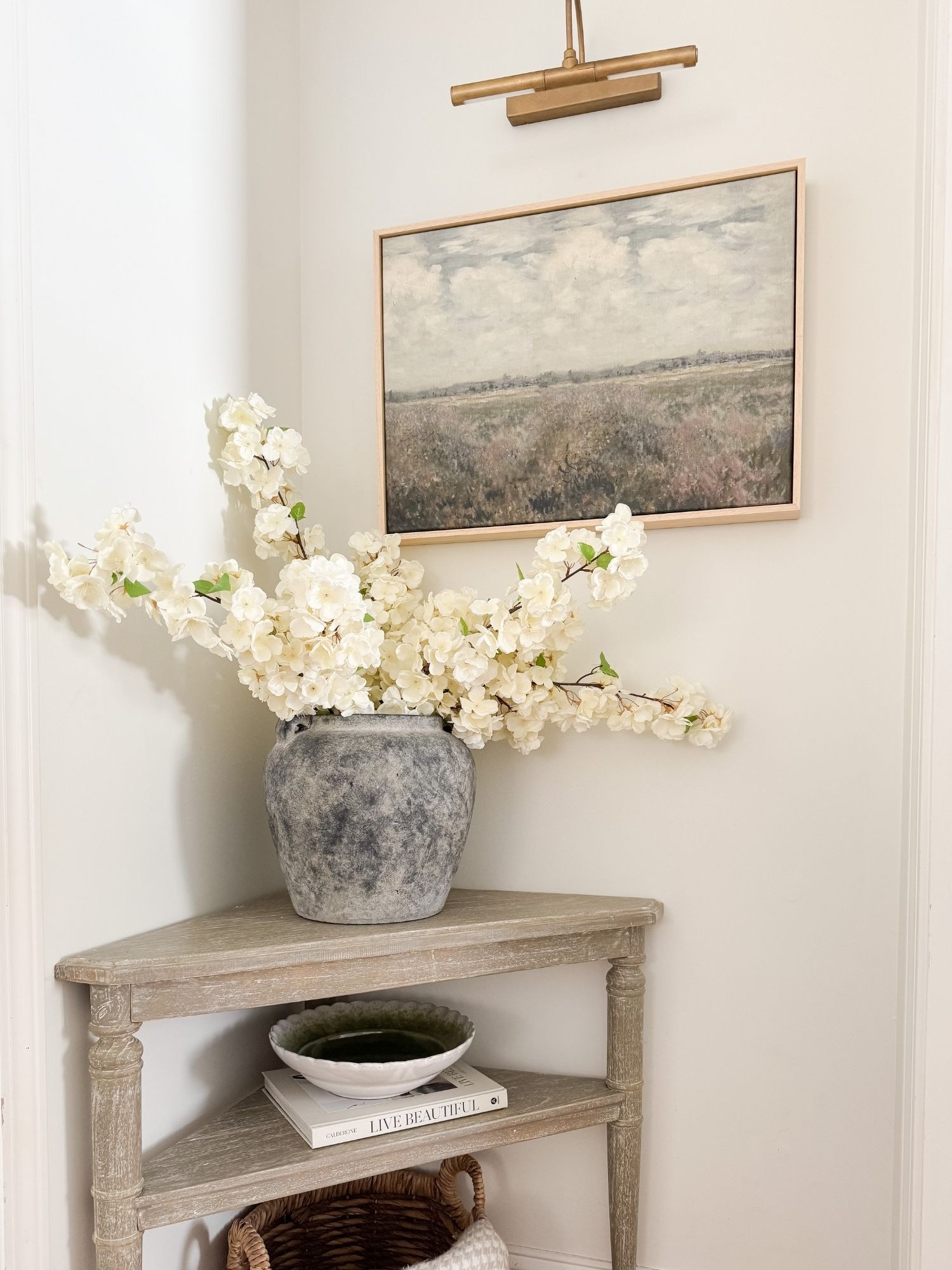 Corner console table styled with textured ceramic vase, white flowering branches, landscape wall art, and brass picture light. Soft neutral tones create a warm, elevated entry or hallway moment. 
console table decor, entry table styling, hallway decor, flowering branches, ceramic vase, landscape wall art, picture light, neutral entryway, coastal entryway decor, spring florals, warm neutrals, classic coastal style, sherri calnan home

#LTKSeasonal #LTKHome