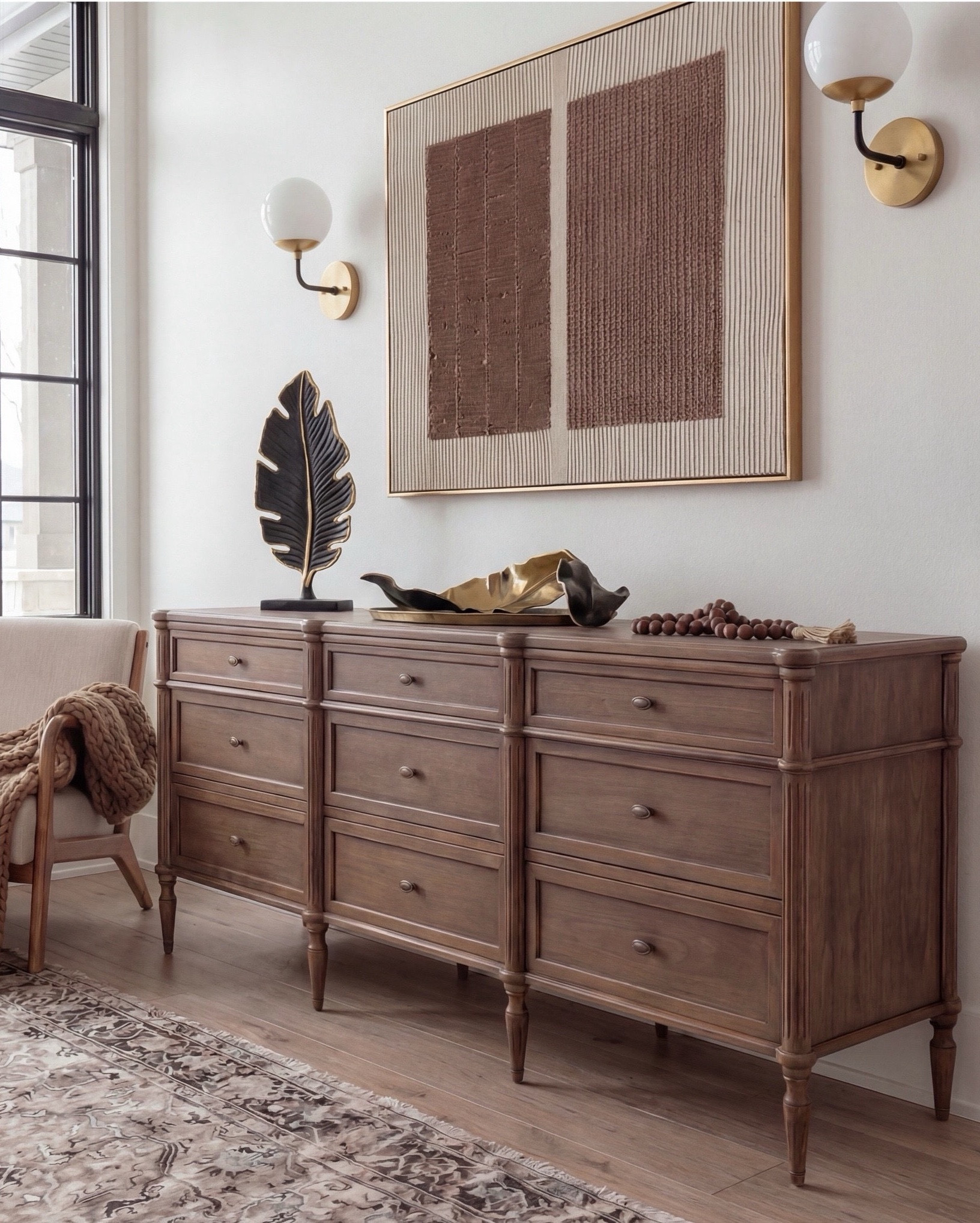 Shop the Look: Bedroom Refresh
• Upholstered bed frame (light neutral fabric)
• White cotton duvet cover + pillow shams
• Neutral patterned area rug
• Chunky knit throw blanket (warm beige)
• Woven knit poufs (foot-of-bed seating)
• Dark wood nightstands with brass pulls
• Sculptural white ceramic table lamps
• White tapered lamp shades
• Brass-framed rectangular mirrors
• Framed floral artwork (warm, moody tones)
• Neutral linen accent pillows
• Ceramic decorative bowl (white sculptural style)
• Small glass vase with white florals
• Dark wood dresser
• Wood upholstered accent chair 
• Brass tray
• Black leaf decor
• Brown wall art

#LTKhome #LTKsale #LTKstyletip