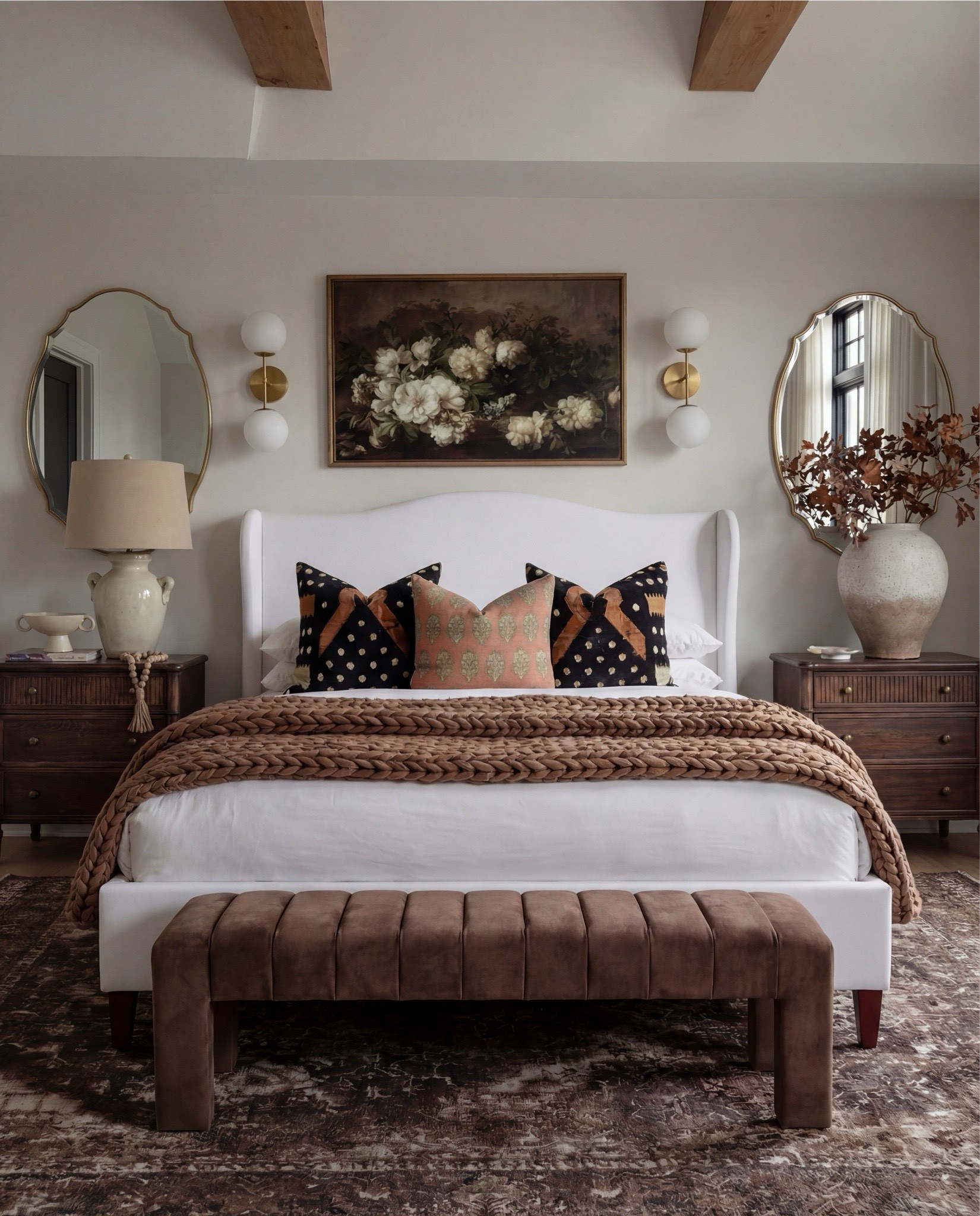 This bedroom feels warm, layered, and quietly refined. The soft upholstered bed keeps the space light and serene, while rich wood tones, earthy textiles, and vintage-inspired accents add depth and contrast. Sculptural lighting, warm brass details, and cozy textures create a balance between elegance and comfort — a space that feels elevated yet deeply livable.

The styling leans into warm neutrals and organic materials — chunky knits, aged woods, soft linen layers, and curated decorative moments that give the room a collected, timeless atmosphere.

Shop the Look:
• White upholstered wingback bed frame
• White cotton duvet cover + pillow shams
• Textured throw pillows (rust + black patterned)
• Chunky knit throw blanket (warm brown)
• Vintage-style brown area rug
• Dark wood nightstands with brass knobs
• Ceramic table lamp (neutral glaze)
• Brass globe wall sconces
• Framed floral artwork (muted tones)
• Arched brass wall mirrors
• Upholstered bench at foot of bed (camel brown)
• Cream accent chair with wood arms
• Black fluted dresser with brass pulls
• Round black fluted end table 
•Fecor books 
• Large ceramic floor vase (earthy clay tone)
• Decorative wood bead garland
• Sheer white curtain panels
•Round decor bowl tray 
•Wood jewellery box 
•Terracotta bowl 

#LTKSaleAlert #LTKHome