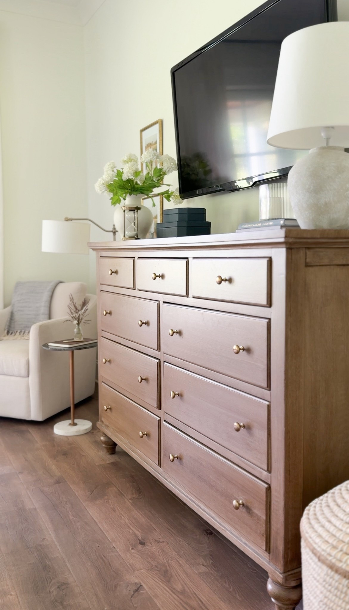 A recent shot of our primary bedroom styled dresser. This dresser was a refinish project inspired by the gorgeous Pottery Barn Sausilito dresser. I painted it to look like wood and changed the hardware. I recently added this curved white vase with artificial snowball hydrangeas I found on Amazon and a set of blue shagreen stacking boxes from Target. To finish off this side of the primary bedroom I added a neutral cozy reading swivel chair and a lamp. 

#LTKHome #LTKSummerEdit