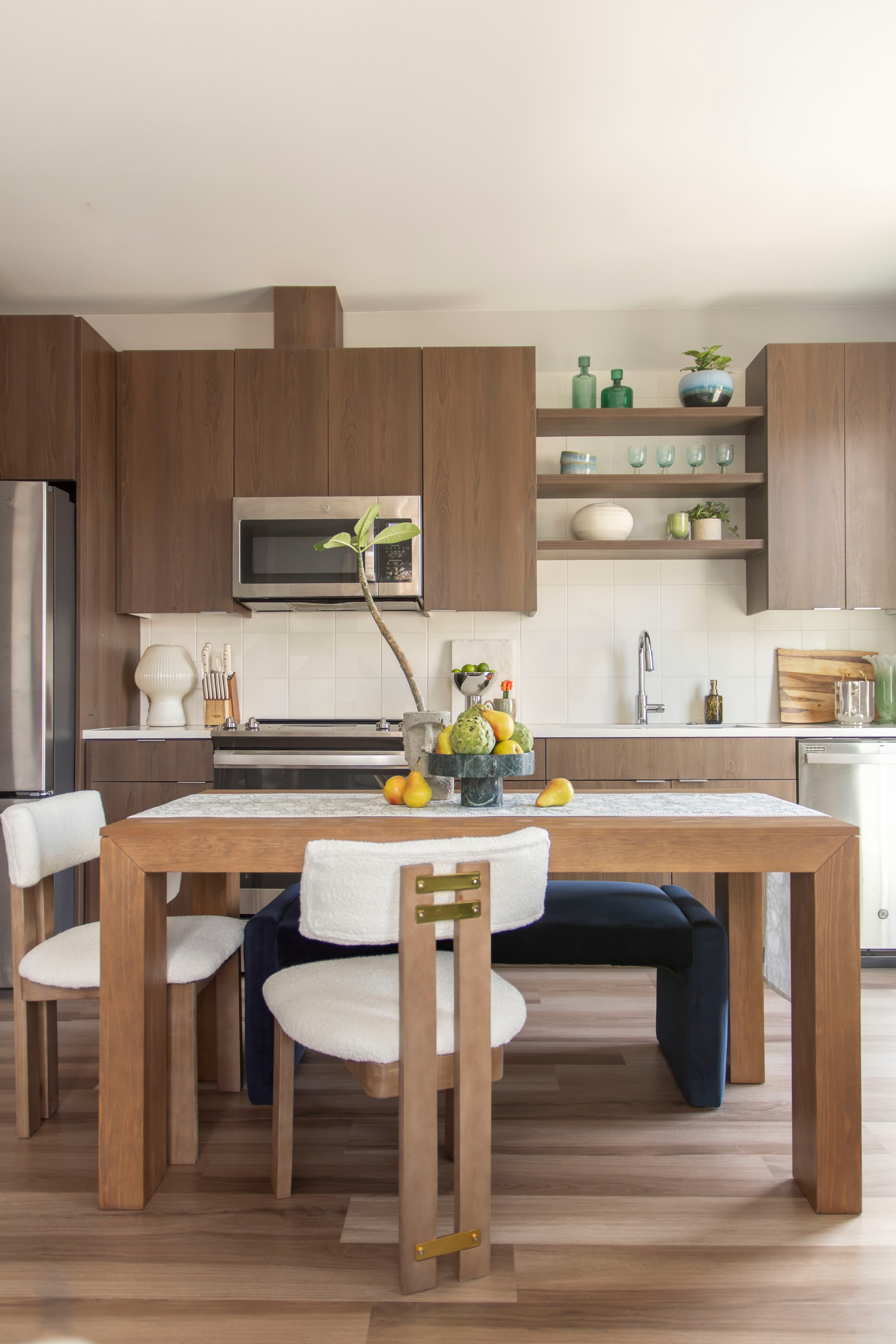 Warm, modern kitchen design with natural wood cabinetry, open shelving, and sleek stainless steel appliances. A contemporary dining table with cozy textured chairs and a minimalist centerpiece creates an inviting space for everyday meals and entertaining. Styled with fresh fruit, neutral tones, and pops of green from plants and glassware, this apartment kitchen blends functionality with chic, organic style. Perfect inspiration for small space living, modern dining setups, and transitional home decor. 

 