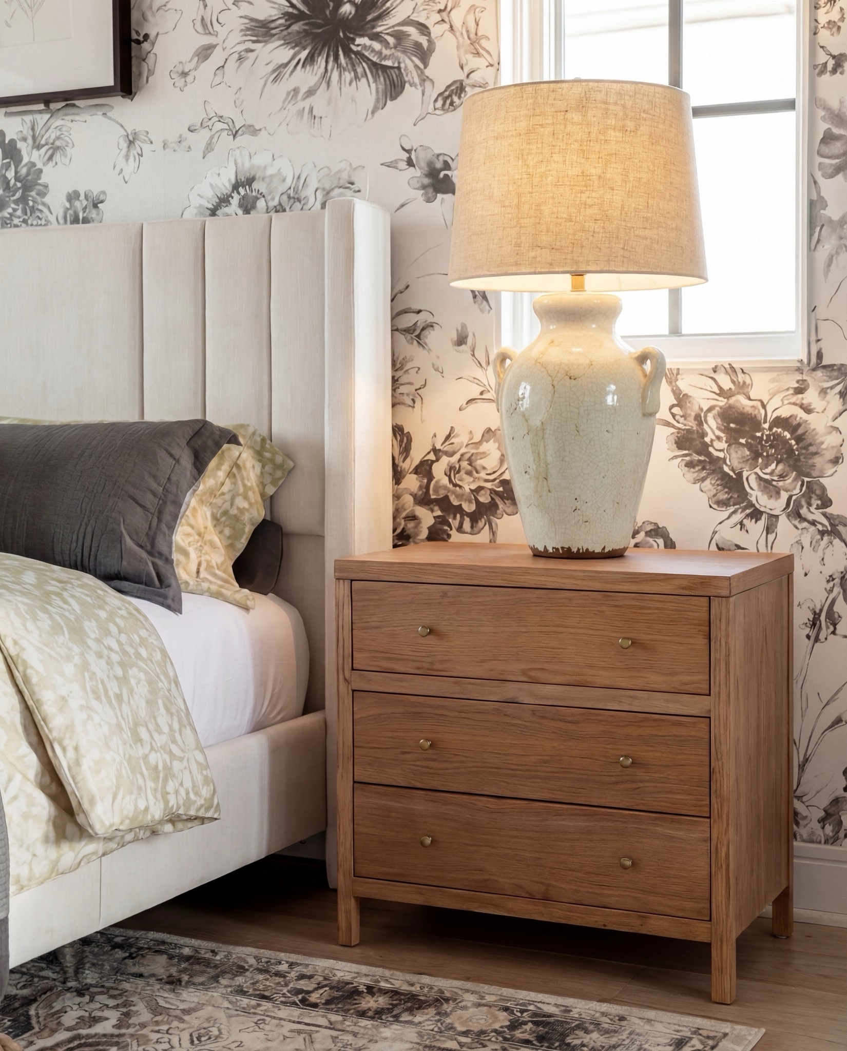Items in this room:
• Upholstered channel-tufted bed — ivory/cream
• Chunky knit throw — warm chestnut brown
• Decorative pillows — muted sage, oatmeal, charcoal
• Light oak nightstands — natural finish
• Ceramic table lamps — soft cream
• Woven ottomans — natural jute
• Patterned area rug — taupe, sand, charcoal
• Accent chair — cream upholstery with warm wood frame
• Botanical wall art — black frames
• Floral wallpaper — charcoal sketch on ivory background

#LTKHome #LTKSaleAlert #LTKSpringSale