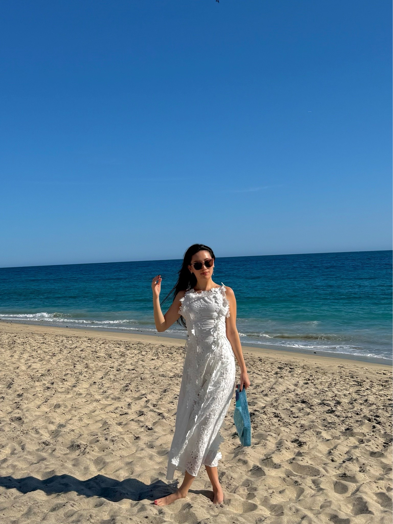 a white floral lace appliqué midi dress with a boat neckline and sleeveless silhouette, styled with tortoiseshell sunglasses and a sky blue woven raffia tote for a barefoot beach day on the baja coast. the dimensional 3d floral lace and clean white silhouette make it the perfect resort dress for cabanas, beach lunches, and sunset walks by the water. perfect for cabo vacations, baja resort getaways, beach club lunches, and mediterranean-inspired coastal trips. shop this look linked above.