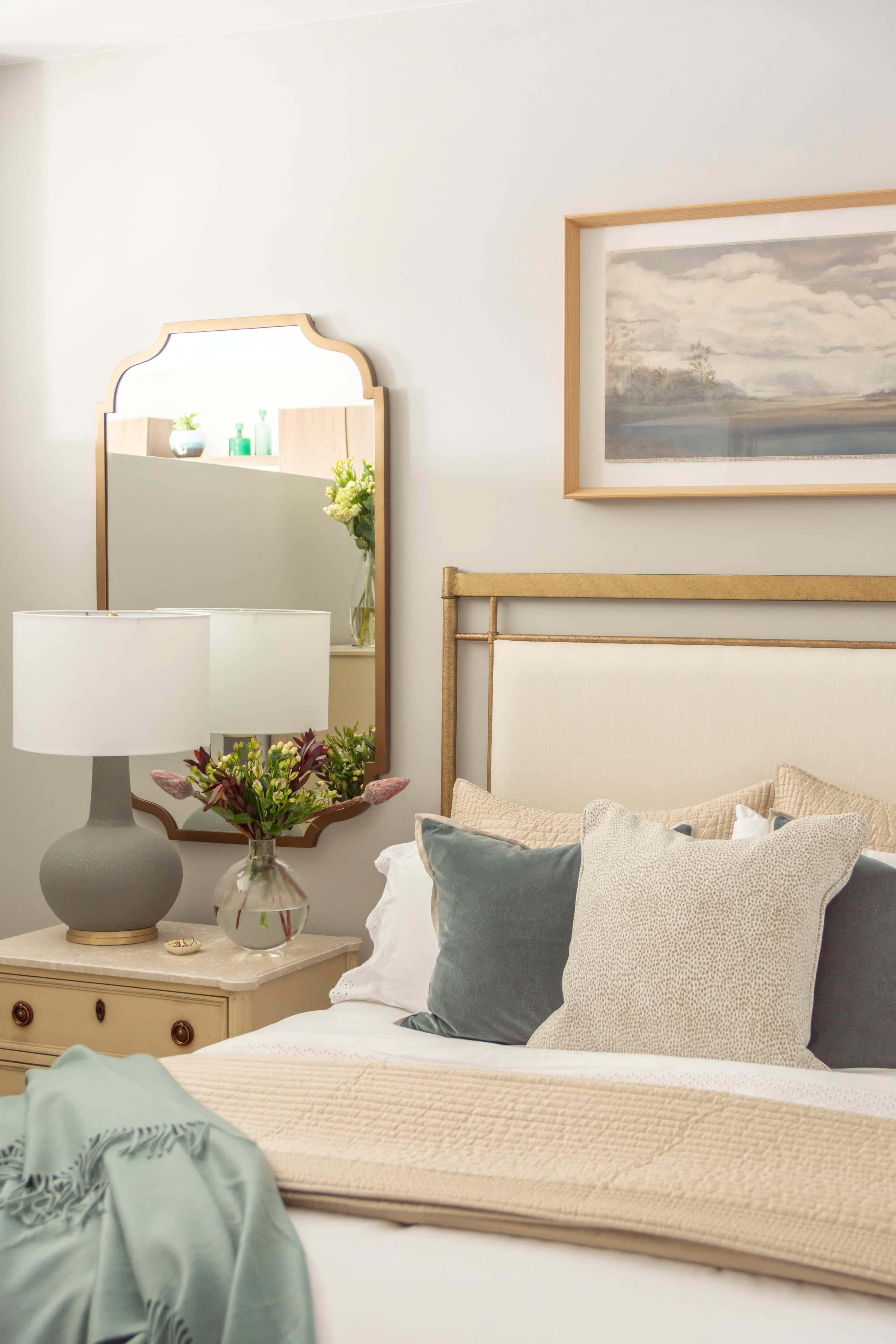 Soft neutrals and cozy textures make this bedroom the perfect retreat. Layered throw pillows in muted blue and cream pair beautifully with a quilted beige coverlet for a timeless look. A vintage-inspired nightstand styled with a round table lamp, glass vase of fresh flowers, and an elegant gold-trimmed mirror adds charm and balance. The gold bed frame and serene landscape artwork complete this airy, modern farmhouse feel. 

 