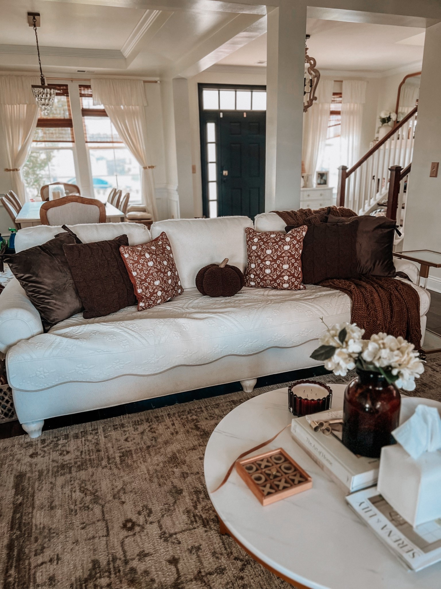 Fall living room
Brown throw pillows
Brown throw blankets
Round marble coffee table 
Brown vase
Brown candle
Cream quilted couch cover 
Brown pumpkin pillow decor 
Gold wooden tic tac toe game
Coffee table book 