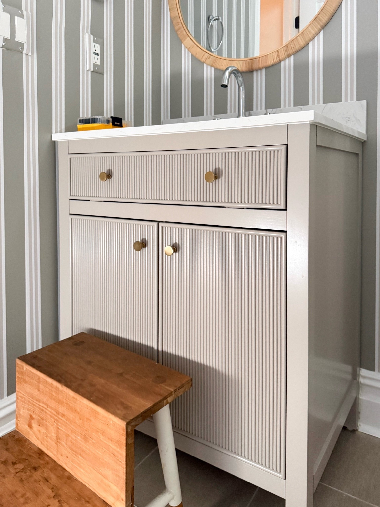 This bathroom vanity is such a beautiful mix of classic and modern details. The fluted cabinet fronts, brass knobs, and warm wood stool add texture and character, while the striped wallpaper ties everything together.

bathroom vanity, fluted bathroom vanity, bathroom cabinet, brass cabinet knobs, striped bathroom wallpaper, bathroom makeover, small bathroom ideas, powder room decor, bathroom storage, modern traditional interiors, bathroom design inspiration, girlonthehudson
