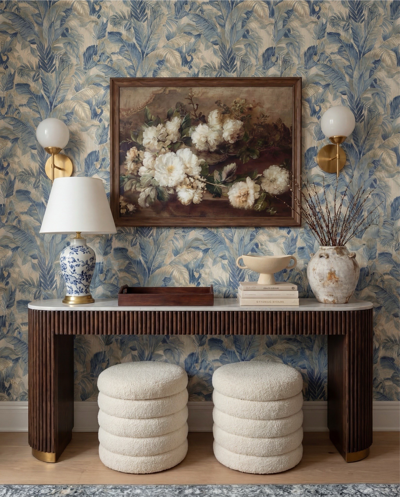 This foyer feels classic, layered, and quietly elegant—the kind of entry that instantly signals a thoughtfully designed home. The blue botanical wallpaper brings softness and movement, while warm wood tones and brass accents ground the space and keep it from feeling too formal. It’s traditional at first glance, but the textures and silhouettes make it feel fresh and livable.

The styling is balanced and intentional—symmetry at the wall, contrast in materials, and sculptural pieces that feel collected over time rather than styled all at once.

Shop the Look:
• Blue botanical wallpaper (leaf motif, muted navy + beige)
• Solid wood console table (dark walnut finish)
• Fluted wood console table legs
• White marble console tabletop
• Framed vintage floral wall art (dark wood frame)
• Brass wall sconces with white globe shades
• Blue and white ceramic table lamp
• White fabric lamp shade (tapered)
• Wooden decorative tray (rectangular)
• Stacked neutral coffee table books
• Sculptural ceramic bowl (cream stoneware)
• Distressed ceramic vase (antique white)
• Dried branches or twigs arrangement
• Bouclé ottomans (ivory, round)
• Light wood flooring
• White baseboards and trim

#LTKHome #LTKSaleAlert