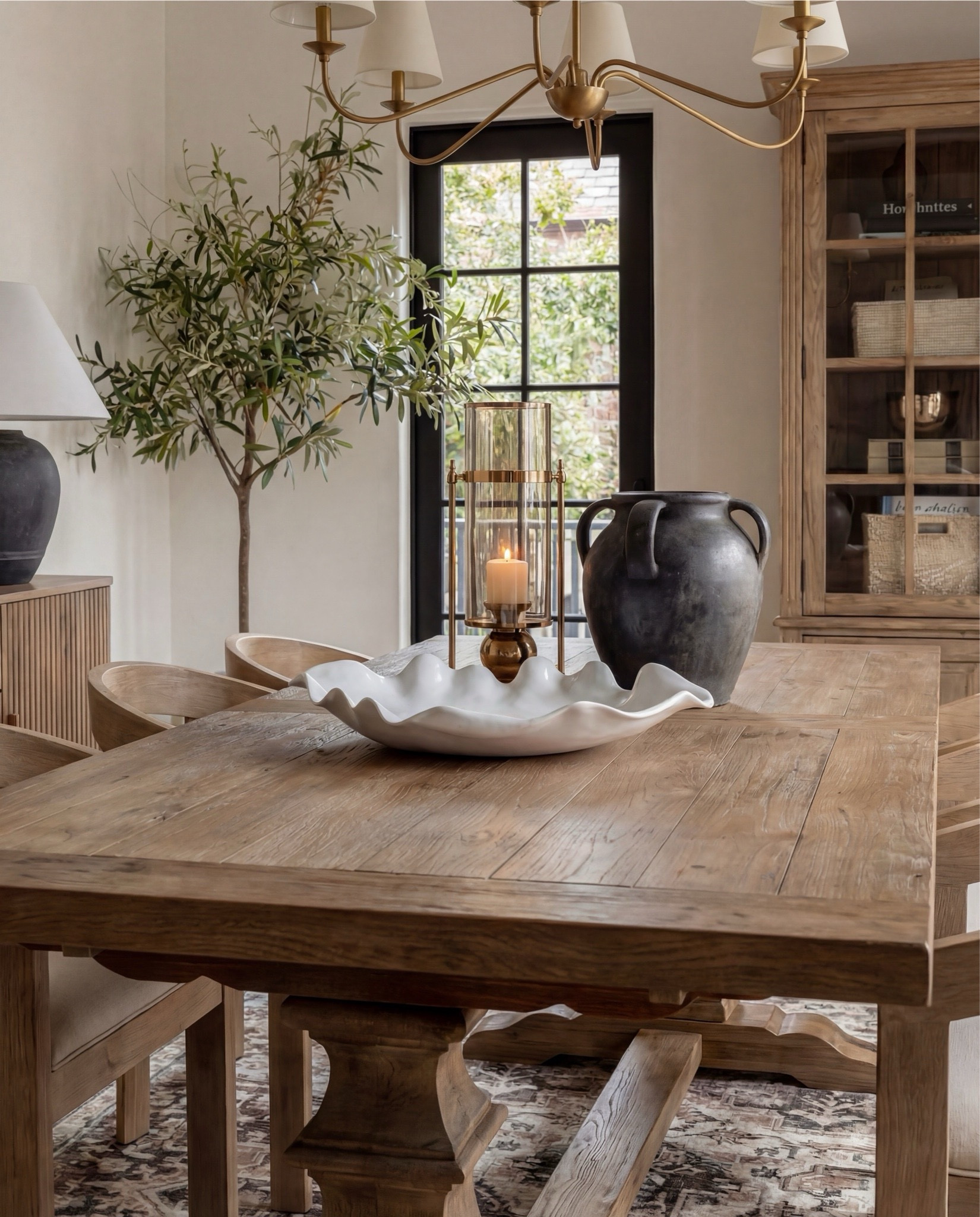 Shop the Look:
• Solid wood pedestal dining table (warm oak tone)
• Light wood dining chairs with upholstered seats
• Neutral vintage-style area rug (soft brown pattern)
• Brass chandelier with tapered fabric shades
• Exposed wood ceiling beams
• Large wood and glass display cabinet
• Fluted wood sideboard cabinet with brass hardware
• Black ceramic table lamp with linen shade
• Framed neutral textile wall art
• Matte black decorative vase
• Wood bead garland with tassels
• Decorative wood tray with ceramic bowl
• Small abstract sculpture decor
• Olive tree in stone planter
• White scalloped ceramic centerpiece bowl
• Dark ceramic pitcher vase (table decor)
• Glass hurricane candle holder
• Sheer neutral curtains

#LTKSaleAlert #LTKHome