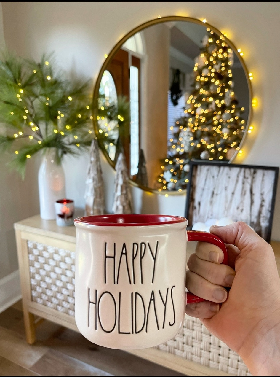 Christmas home decor in the living room. Christmas tree, Christmas garland, pine branches, large round mirror, woven console table, Christmas lights, Happy Holidays Rae Dunn Mug, home decor. 

#LTKHoliday #LTKhome #LTKSeasonal