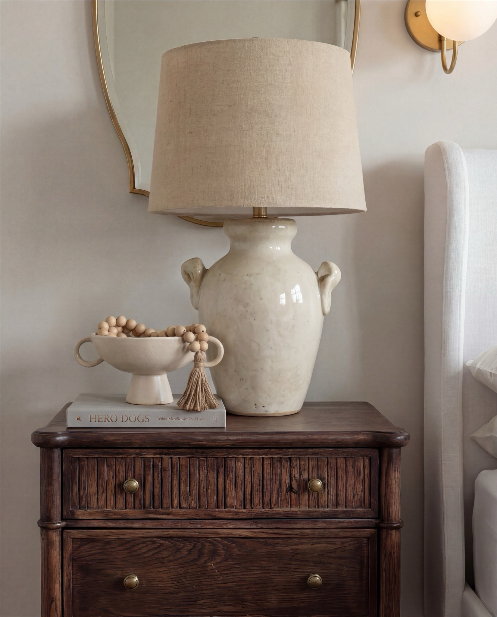 This bedroom feels warm, layered, and quietly refined. The soft upholstered bed keeps the space light and serene, while rich wood tones, earthy textiles, and vintage-inspired accents add depth and contrast. Sculptural lighting, warm brass details, and cozy textures create a balance between elegance and comfort — a space that feels elevated yet deeply livable.

The styling leans into warm neutrals and organic materials — chunky knits, aged woods, soft linen layers, and curated decorative moments that give the room a collected, timeless atmosphere.

Shop the Look:
• White upholstered wingback bed frame
• White cotton duvet cover + pillow shams
• Textured throw pillows (rust + black patterned)
• Chunky knit throw blanket (warm brown)
• Vintage-style brown area rug
• Dark wood nightstands with brass knobs
• Ceramic table lamp (neutral glaze)
• Brass globe wall sconces
• Framed floral artwork (muted tones)
• Arched brass wall mirrors
• Upholstered bench at foot of bed (camel brown)
• Cream accent chair with wood arms
• Black fluted dresser with brass pulls
• Round black fluted end table 
•Fecor books 
• Large ceramic floor vase (earthy clay tone)
• Decorative wood bead garland
• Sheer white curtain panels
•Round decor bowl tray 
•Wood jewellery box 
•Terracotta bowl 

#LTKSaleAlert #LTKHome