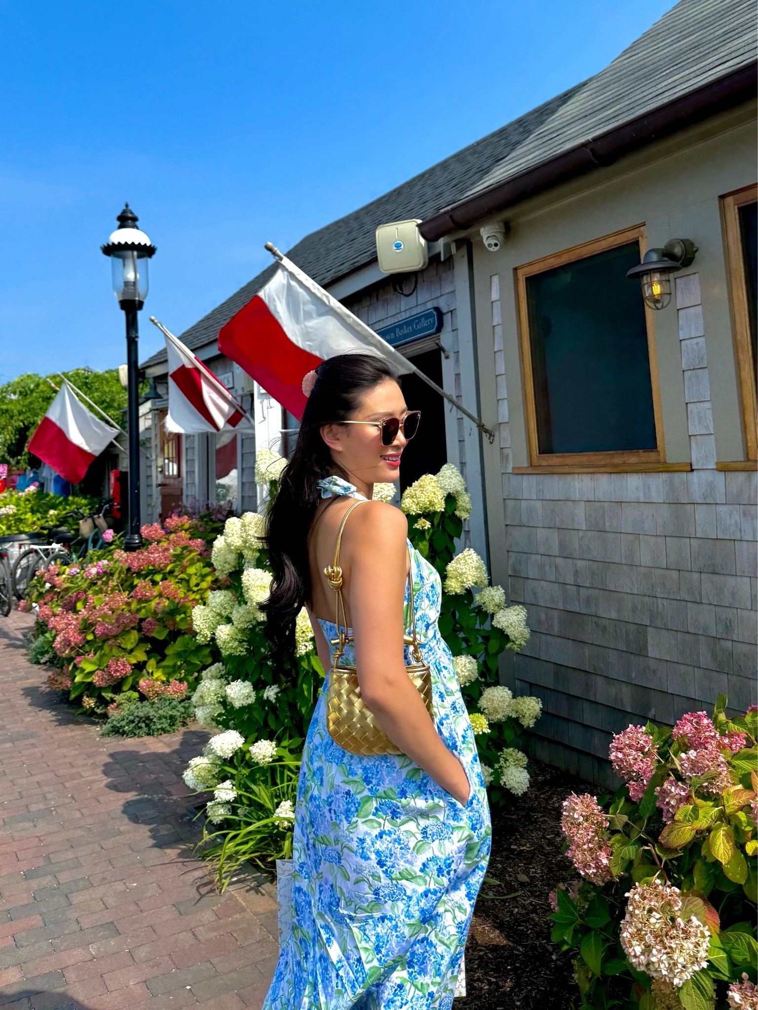 a blue hydrangea floral print maxi dress with a fitted bodice, open back, and tie straps, styled with a gold woven mini shoulder bag, tortoiseshell sunglasses, and a pink ribbon hair tie for an afternoon strolling the cobblestone streets of nantucket old town. the soft watercolor hydrangea print and feminine open-back silhouette make this the ultimate young old money nantucket summer dress. perfect for nantucket trips, hamptons summer afternoons, martha’s vineyard getaways, and coastal new england vacations. shop this look linked above.