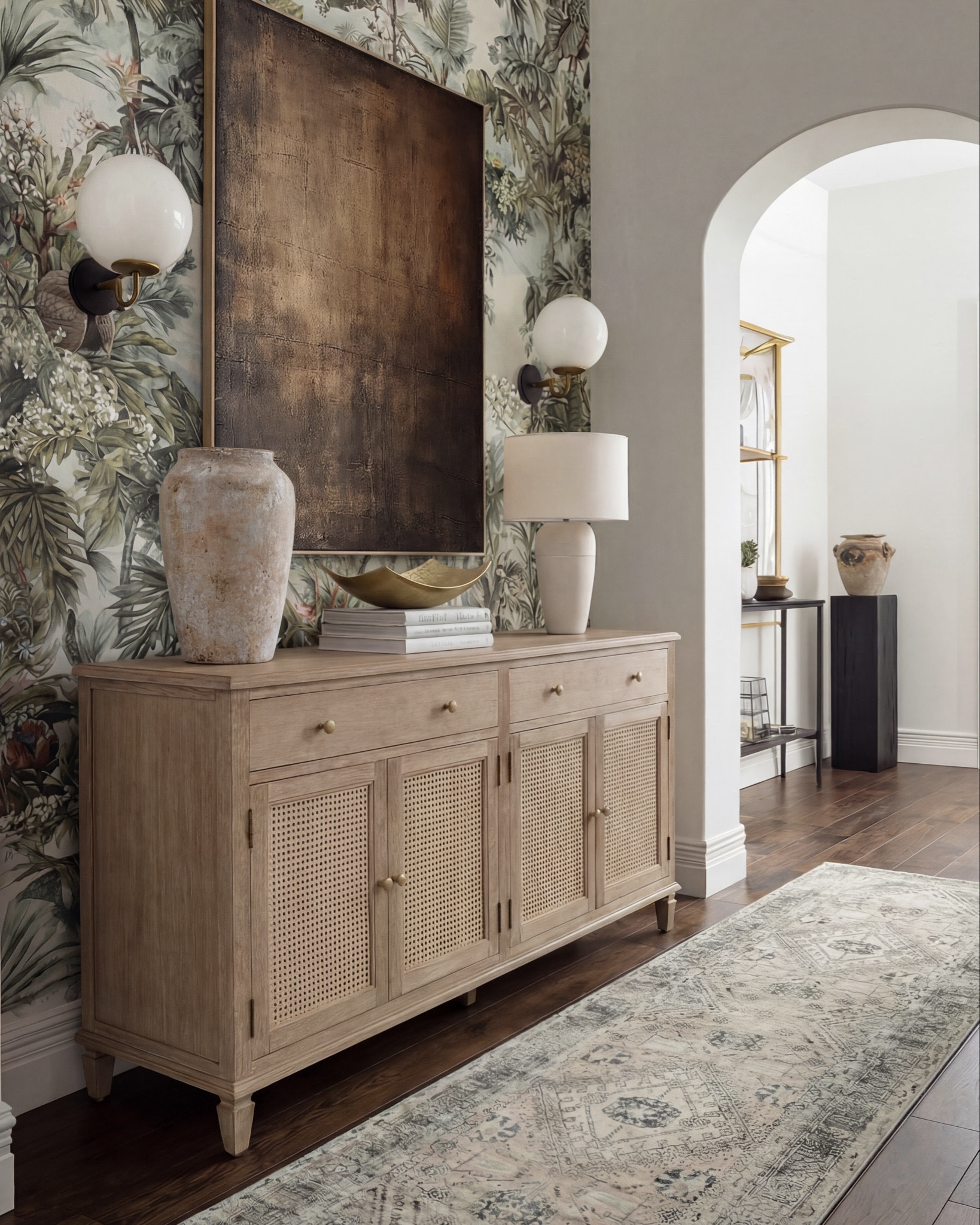 There’s something about a moody, nature-inspired foyer that just sets the tone for the whole home, and this one does it so effortlessly. The mix of warm wood, soft texture, and bold art makes the space feel collected and elevated from the moment you walk in.

The natural-oak console brings the perfect balance of clean lines and subtle detail with its woven cane doors. I styled it with an oversized stone vase, a tall ceramic lamp, and a sculptural gold bowl layered on books — just enough to feel intentional without being cluttered.

The vintage-inspired runner adds muted pattern and softness underfoot, grounding all the warm wood tones beautifully. And that wallpaper — the deep greens, palms, and floral textures make the entire wall feel like a piece of art on its own.

The oversized copper-toned artwork brings so much richness to the scene, pulling in the darker notes from the sconces and the natural shadows in the wallpaper. It’s the kind of statement piece that instantly elevates a foyer.

✨ Shop the Look
Natural-oak cane console | Oversized stone vase | Sculptural gold bowl | Ceramic table lamp | Botanical wallpaper | Vintage-style runner | Copper-toned oversized artwork | Globe wall sconces

#LTKsale #LTKhome #LTKstyletip